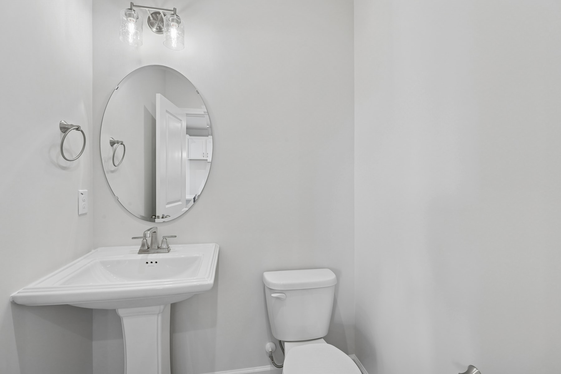 A minimalist bathroom with a white pedestal sink, a round mirror, and a toilet against a plain white wall.