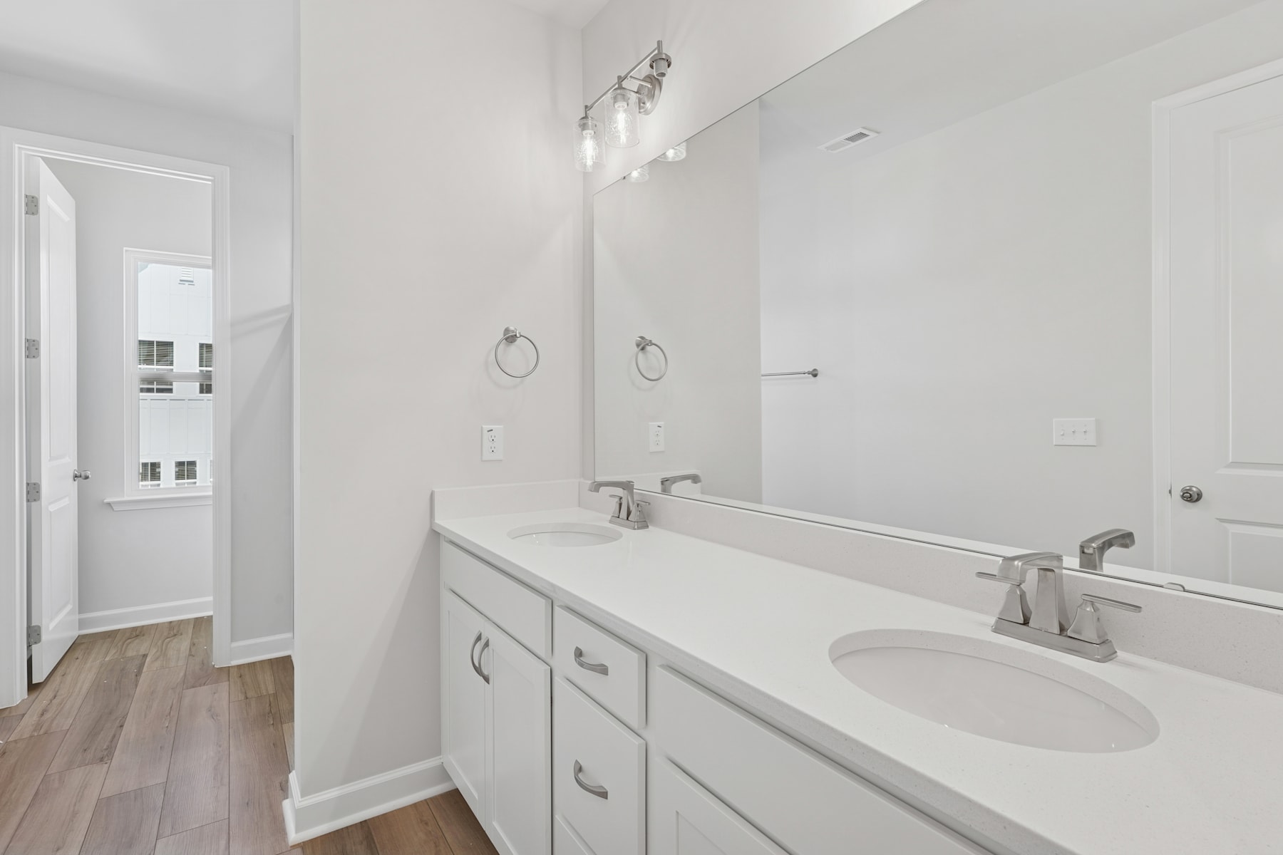 A modern, well-lit bathroom with a white vanity, sink, and mirror, set against a neutral-toned wall and hardwood flooring.