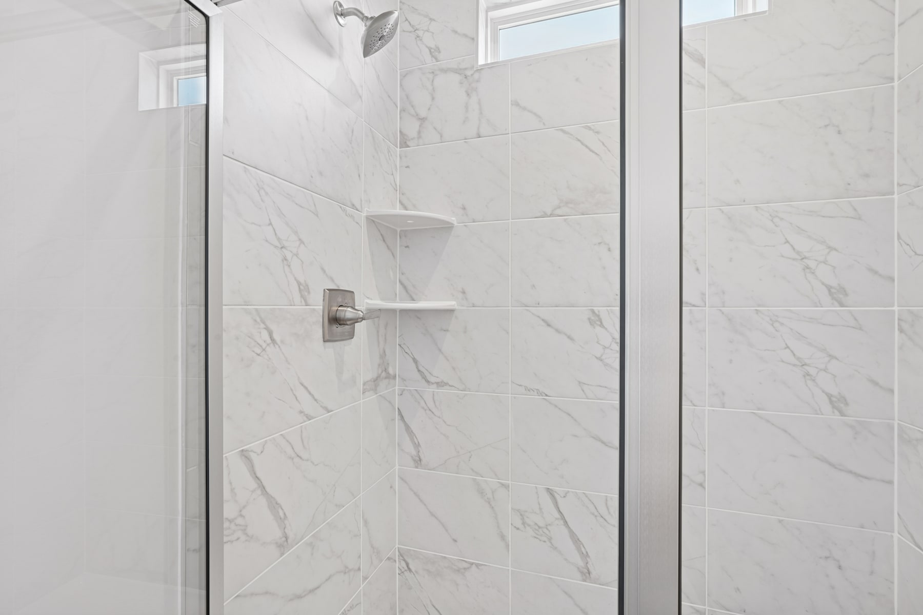 A modern, minimalist bathroom with a glass shower enclosure and white marble-like tiles covering the walls and floor.