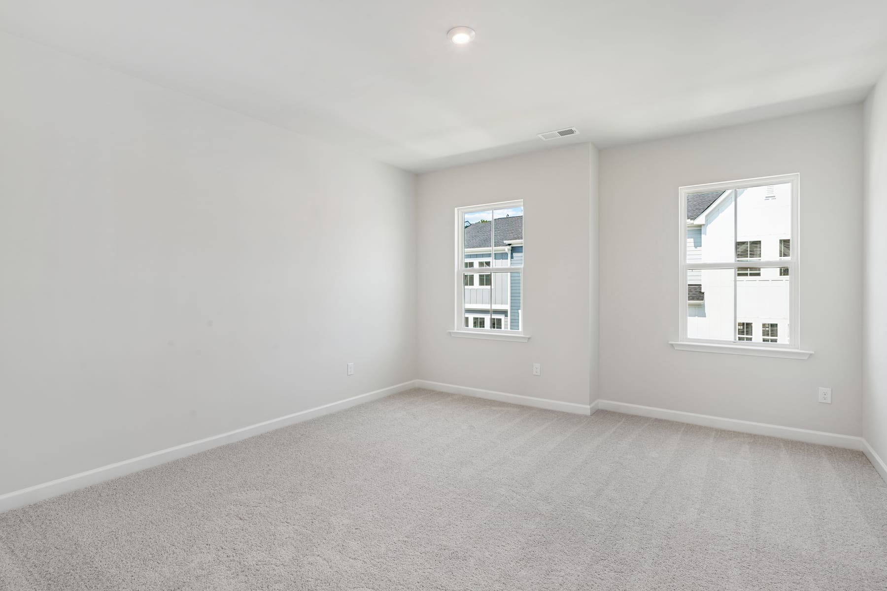 A bright, empty room with white walls, a light-colored carpet, and large windows providing natural light.