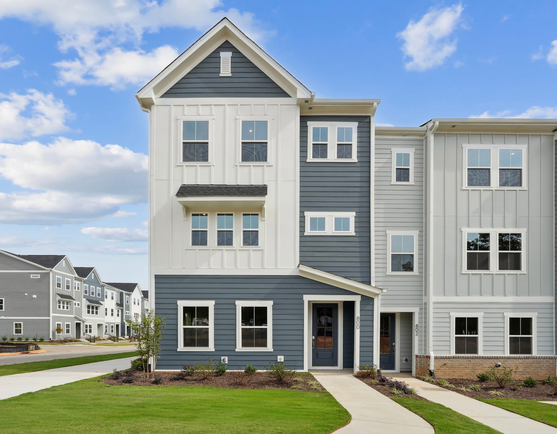 A multi-story residential building with a gray exterior and white trim, surrounded by a well-manicured lawn and set against a blue sky with scattered clouds.