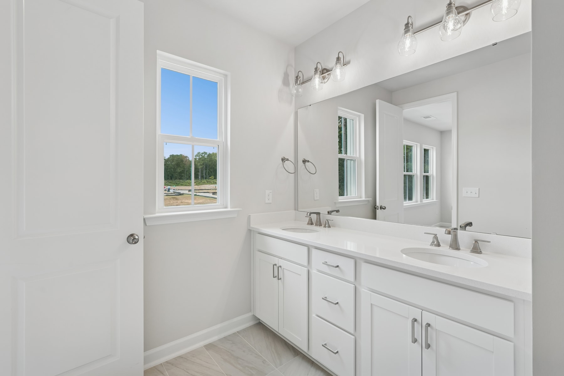 A bright and spacious bathroom with white cabinets, a large mirror, and a window overlooking a scenic outdoor view.