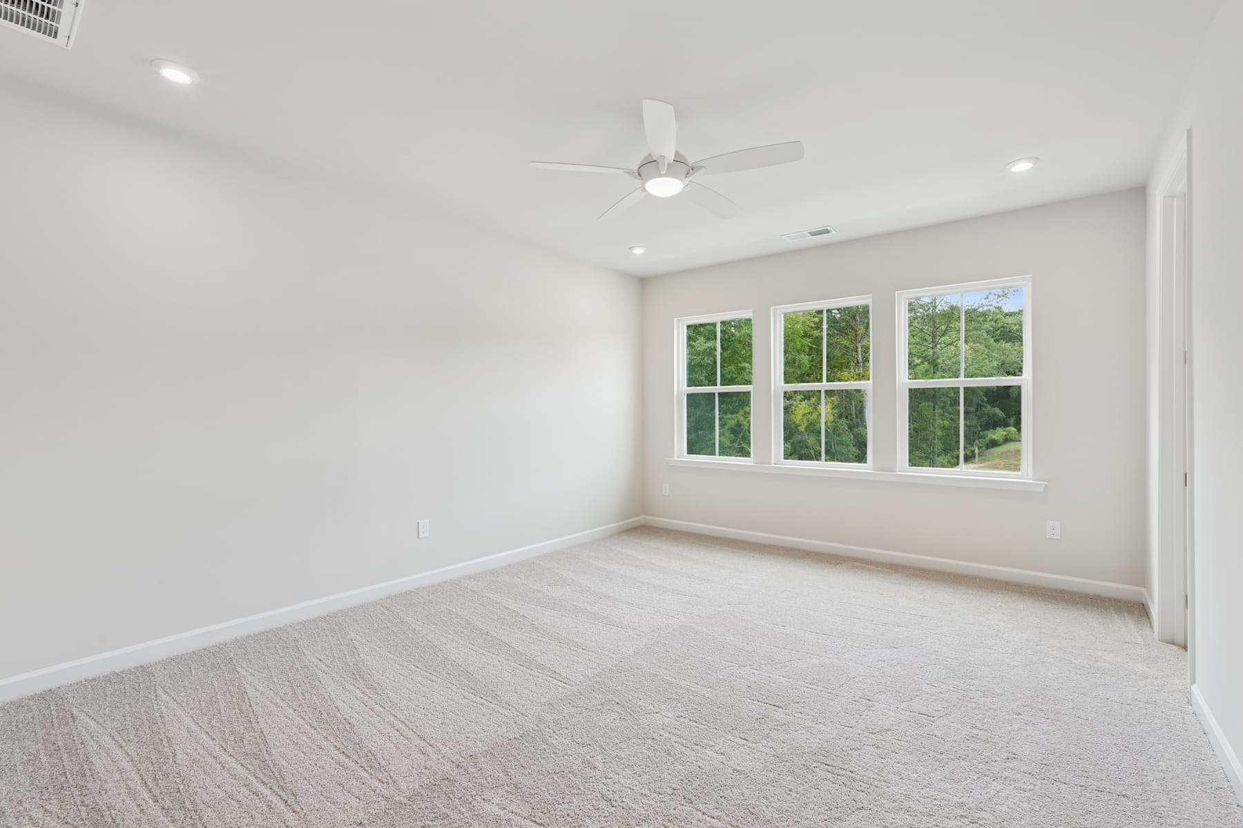 A bright, spacious room with large windows overlooking a lush, green outdoor area, featuring a ceiling fan and a neutral-toned carpet flooring.