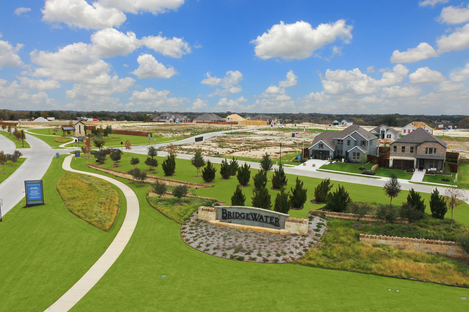 A well-manicured residential community with lush green lawns, neatly landscaped gardens, and a winding path leading through the development, set against a backdrop of a cloudy blue sky.