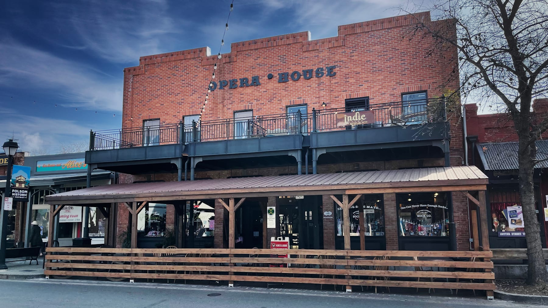 A brick building with the words "Opera House" prominently displayed, surrounded by a wooden deck and benches, set against a blue sky with some trees in the background.