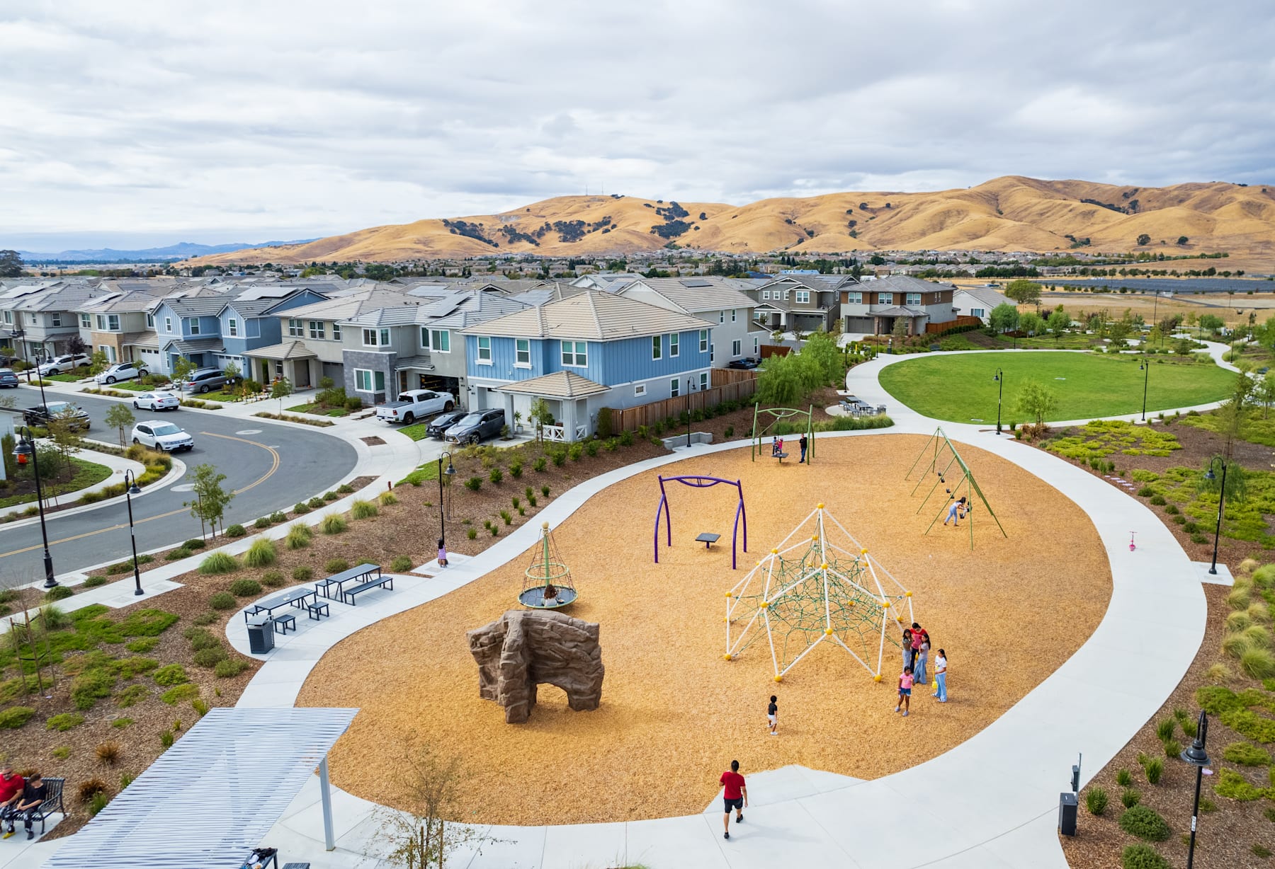 A residential neighborhood with colorful townhouses, a playground with swings and a large sculpture in the foreground, surrounded by rolling hills and a cloudy sky in the background.