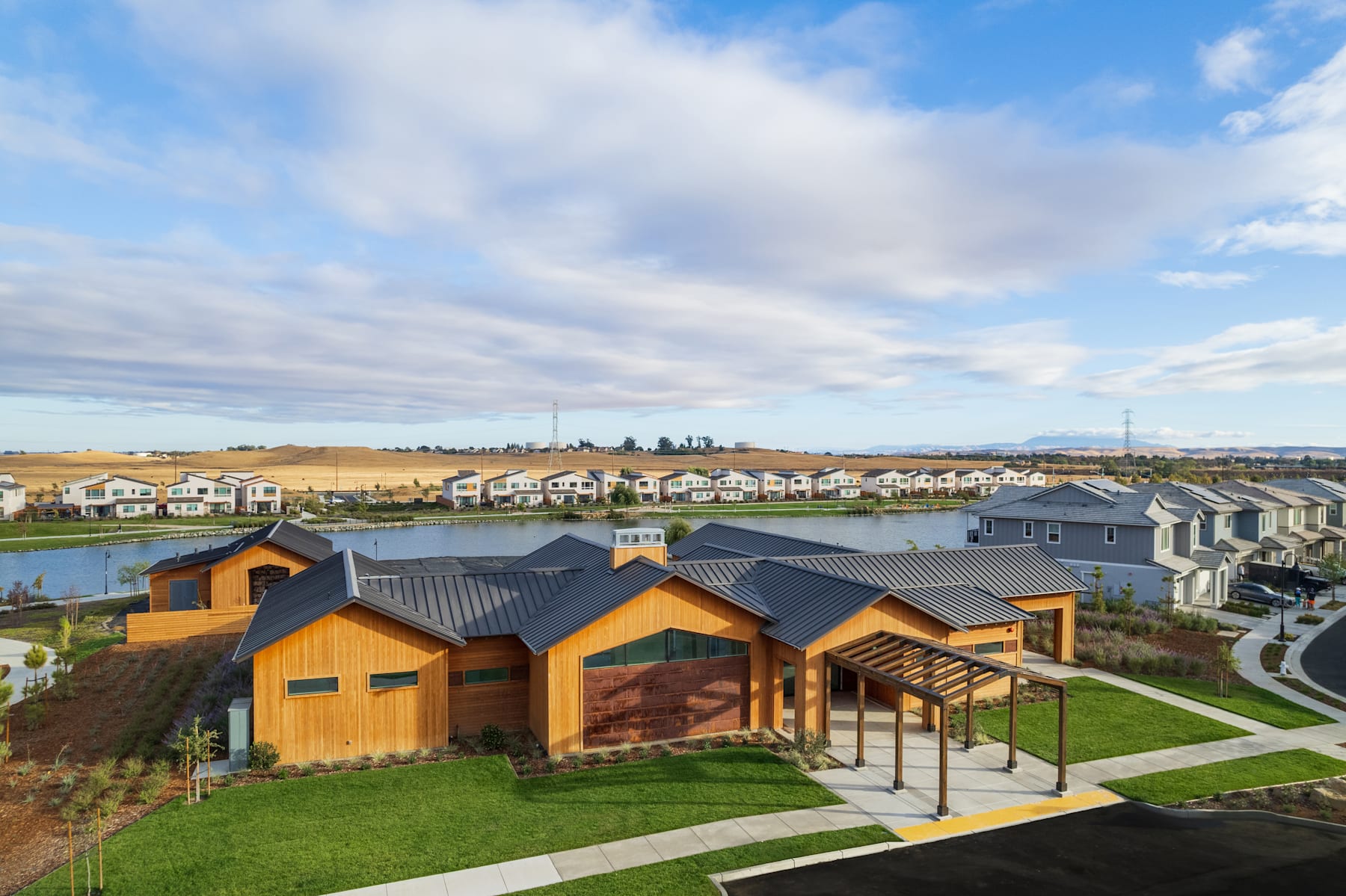 A residential neighborhood with colorful wooden houses surrounded by lush green lawns, set against a backdrop of a cloudy blue sky.