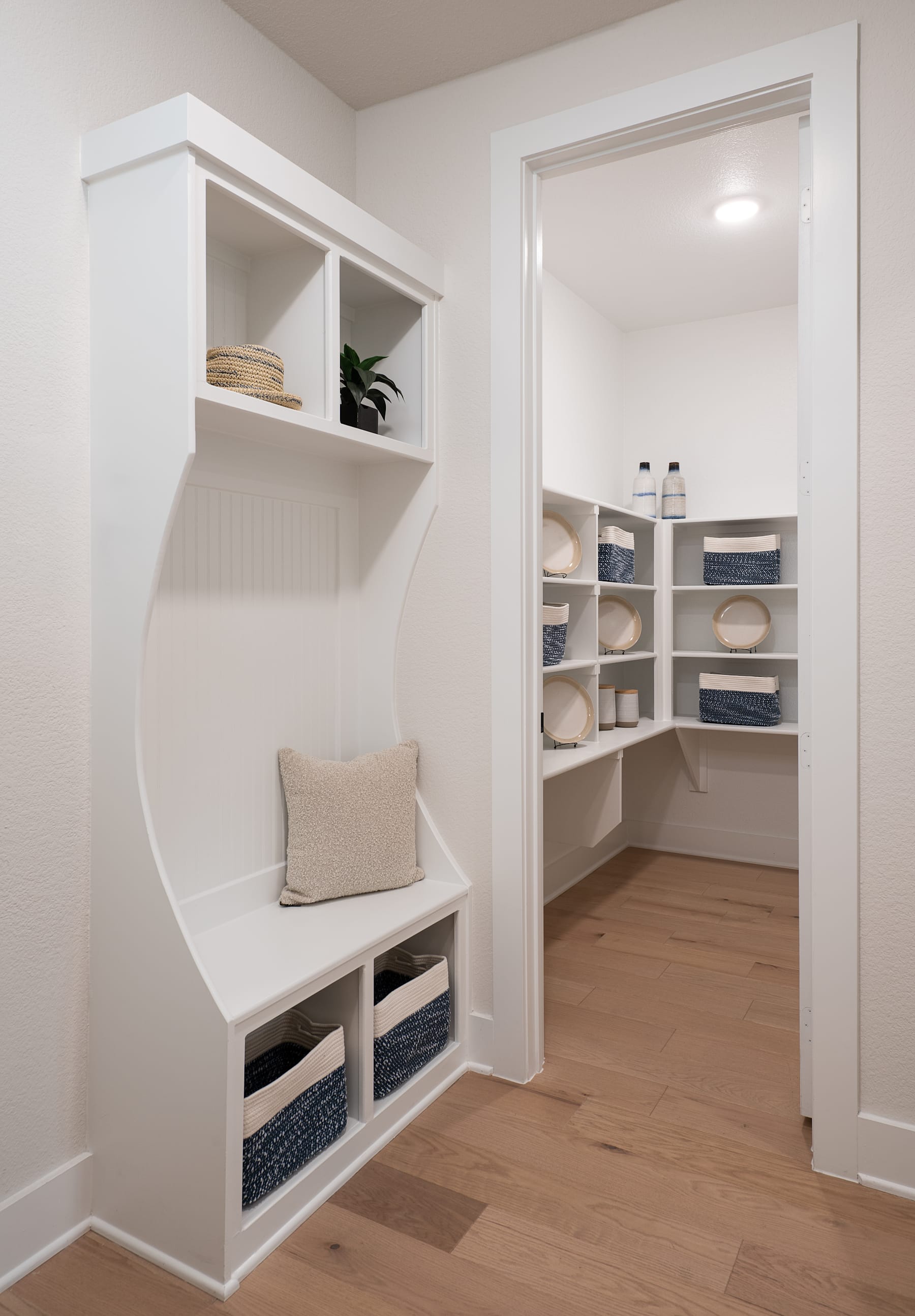 A well-organized and minimalist mudroom or entryway with built-in shelving, storage cubbies, and a bench, leading into a closet or pantry space with additional shelving.