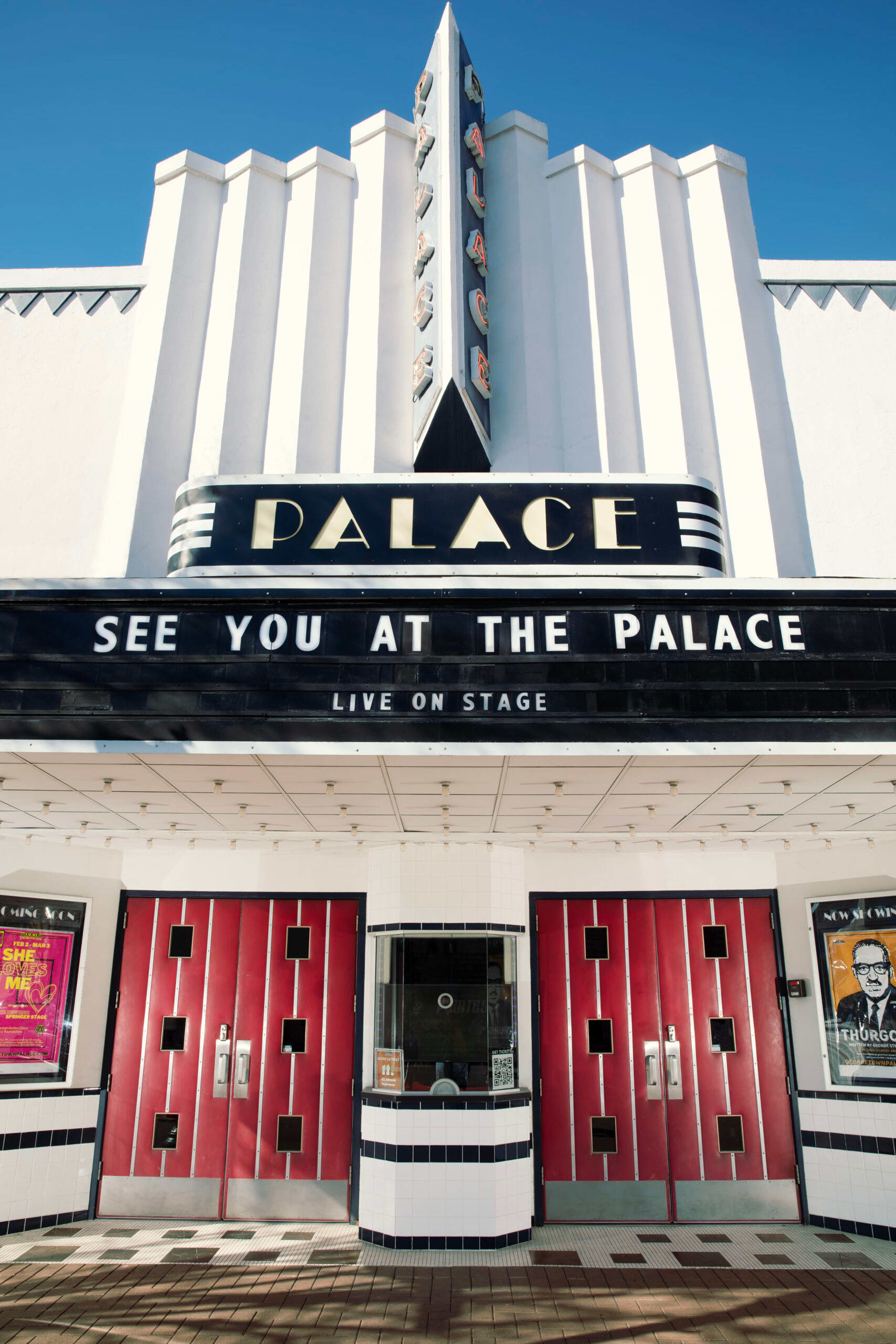 A large, white, art deco-style building with the word "PALACE" prominently displayed on the marquee, which reads "SEE YOU AT THE PALACE LIVE ON STAGE", with red doors in the foreground.