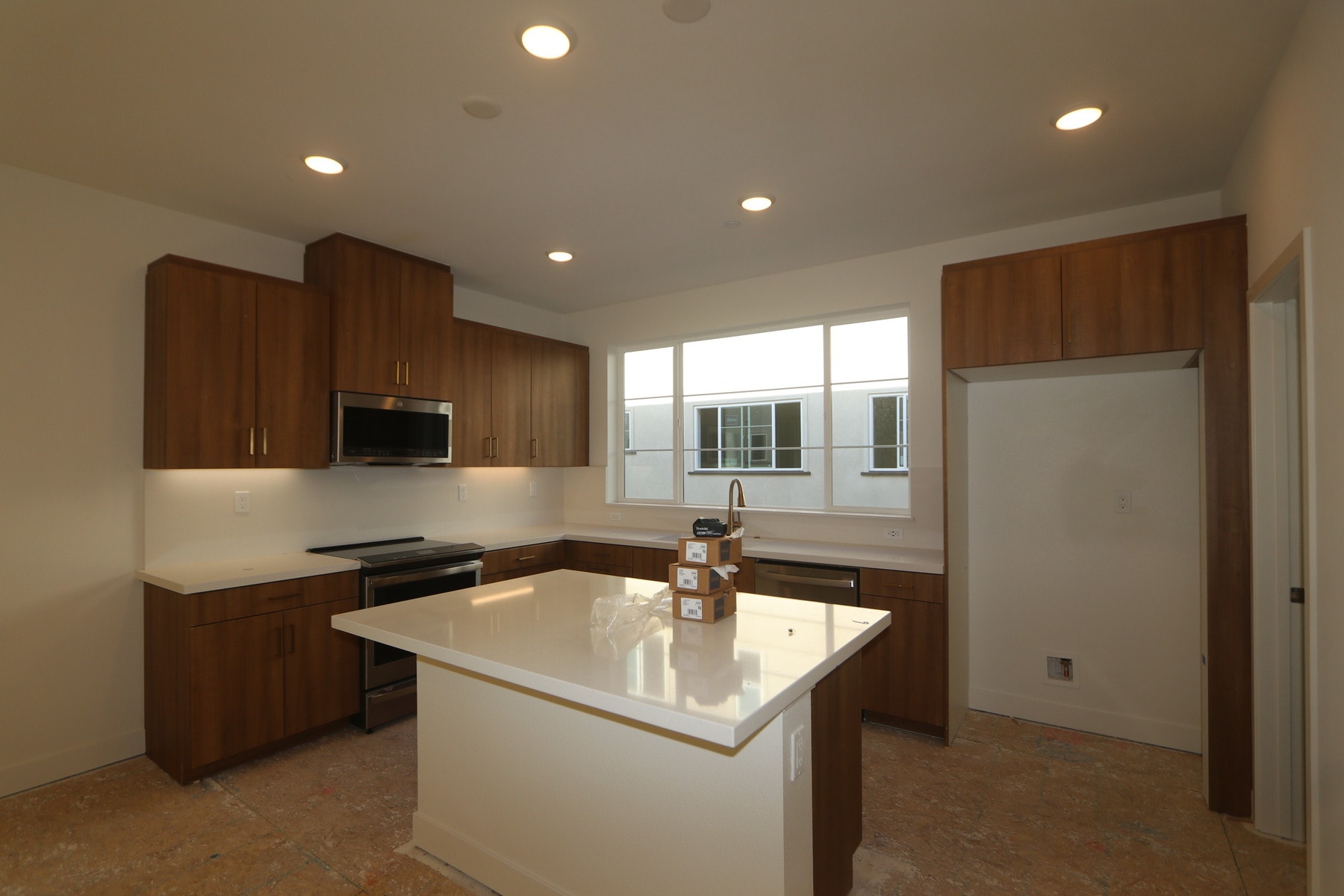 A modern kitchen with wooden cabinets, a white countertop, and recessed lighting, set against a bright window in the background.