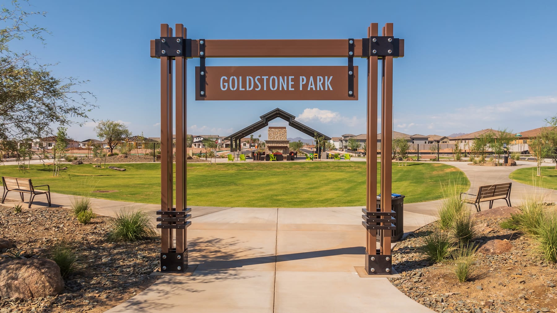 A wooden sign with the text "Goldstone Park" stands at the entrance of a well-maintained park, surrounded by lush greenery and residential buildings in the background.