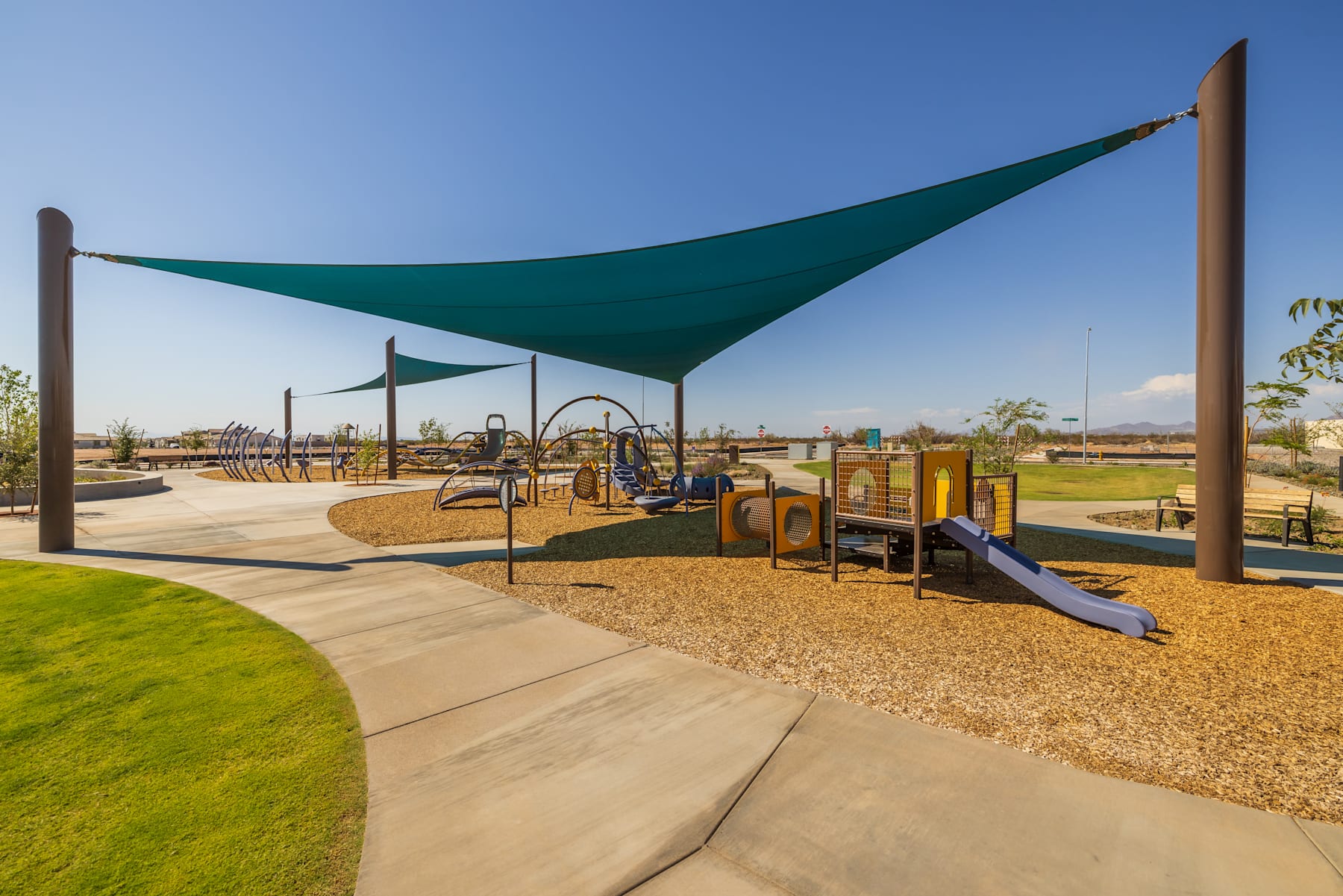 A shaded playground area with various play equipment, surrounded by a grassy landscape and a clear blue sky.