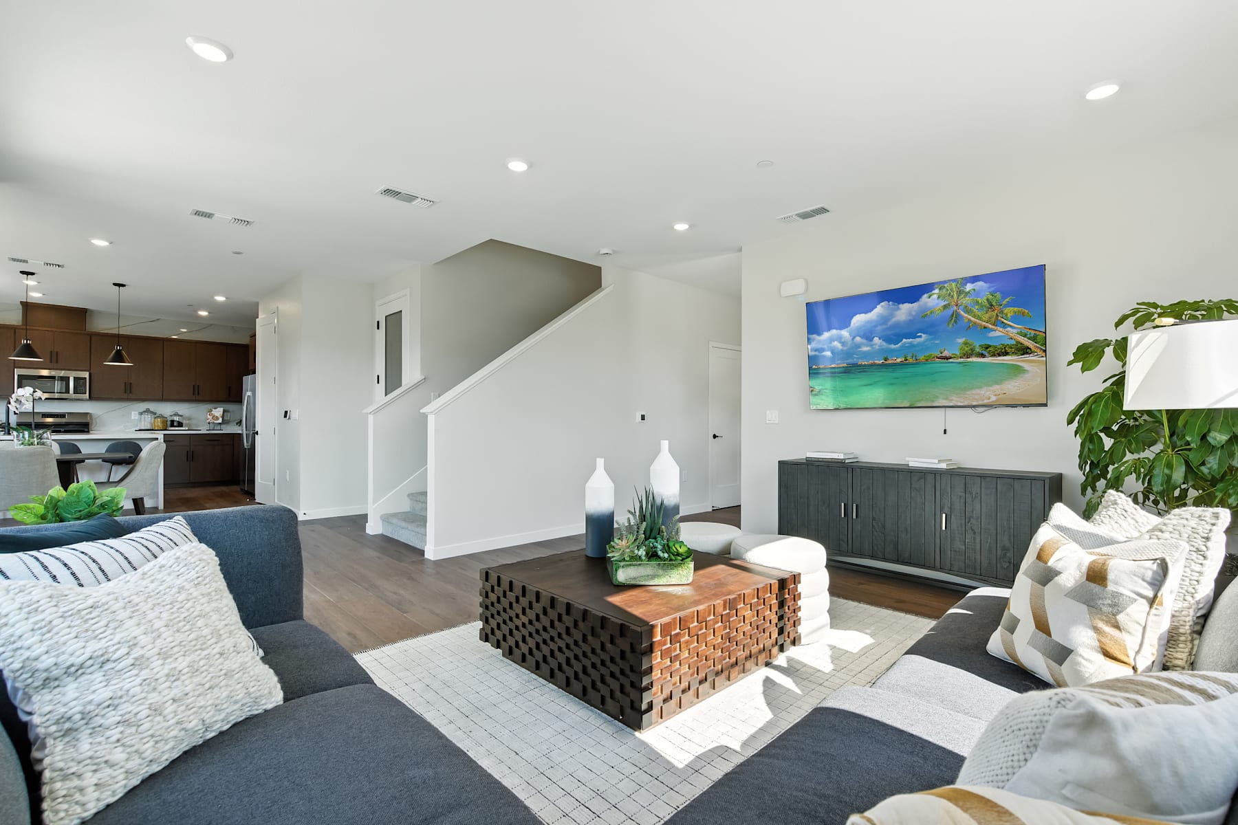 A modern and cozy living room with a gray sectional sofa, a woven coffee table, and a vibrant artwork depicting a tropical beach scene on the wall.