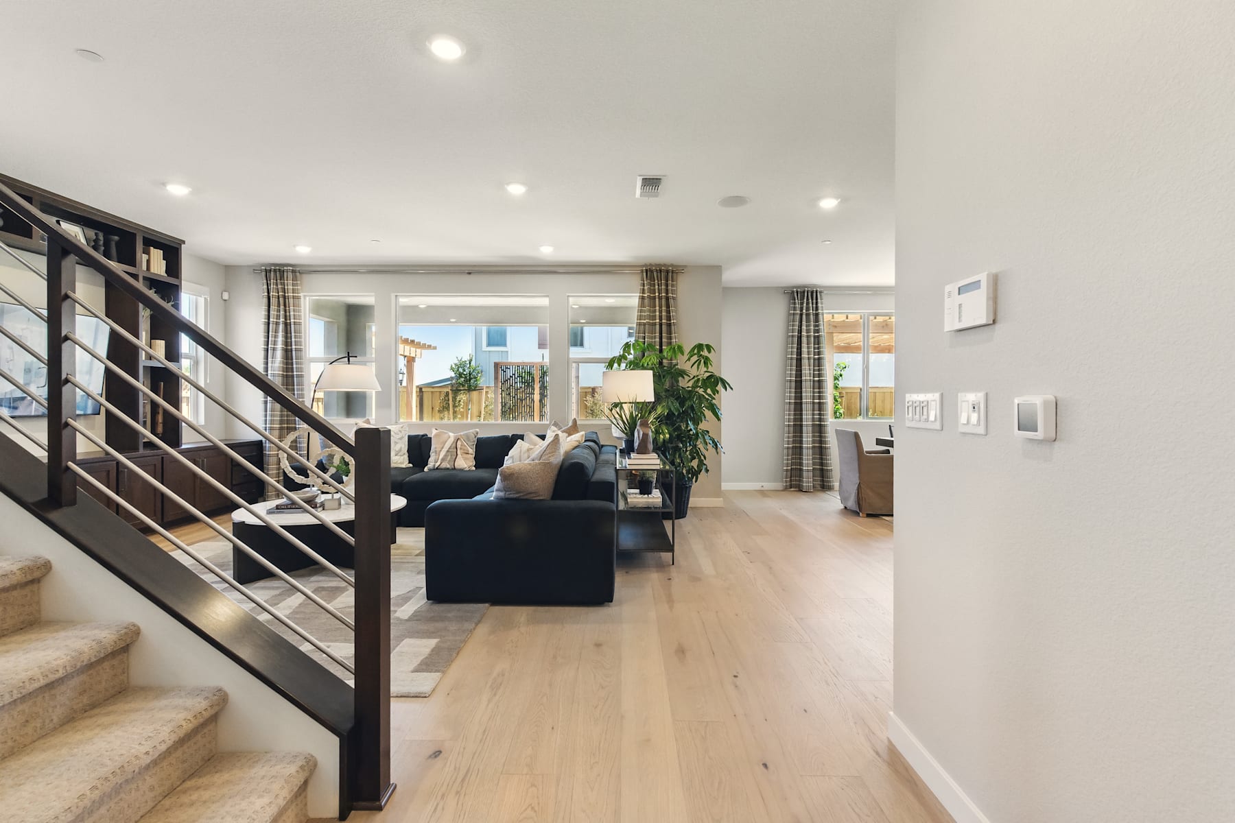 A modern and spacious living room with a staircase leading to the upper level, featuring hardwood floors, a black sofa, and a potted plant in the background.