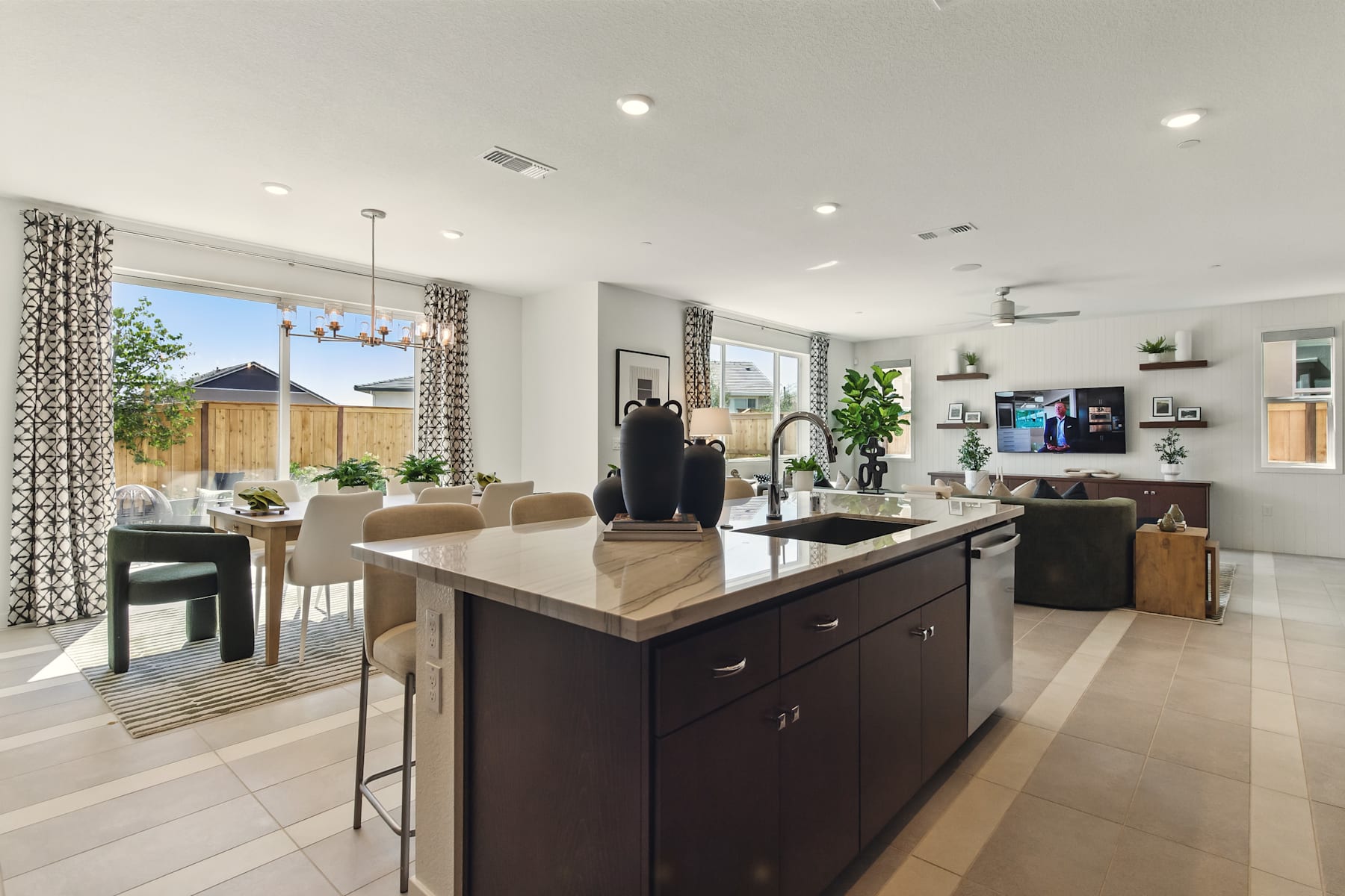 A modern and spacious kitchen with a large island, dark cabinets, and a dining area visible in the background, surrounded by large windows providing natural light.