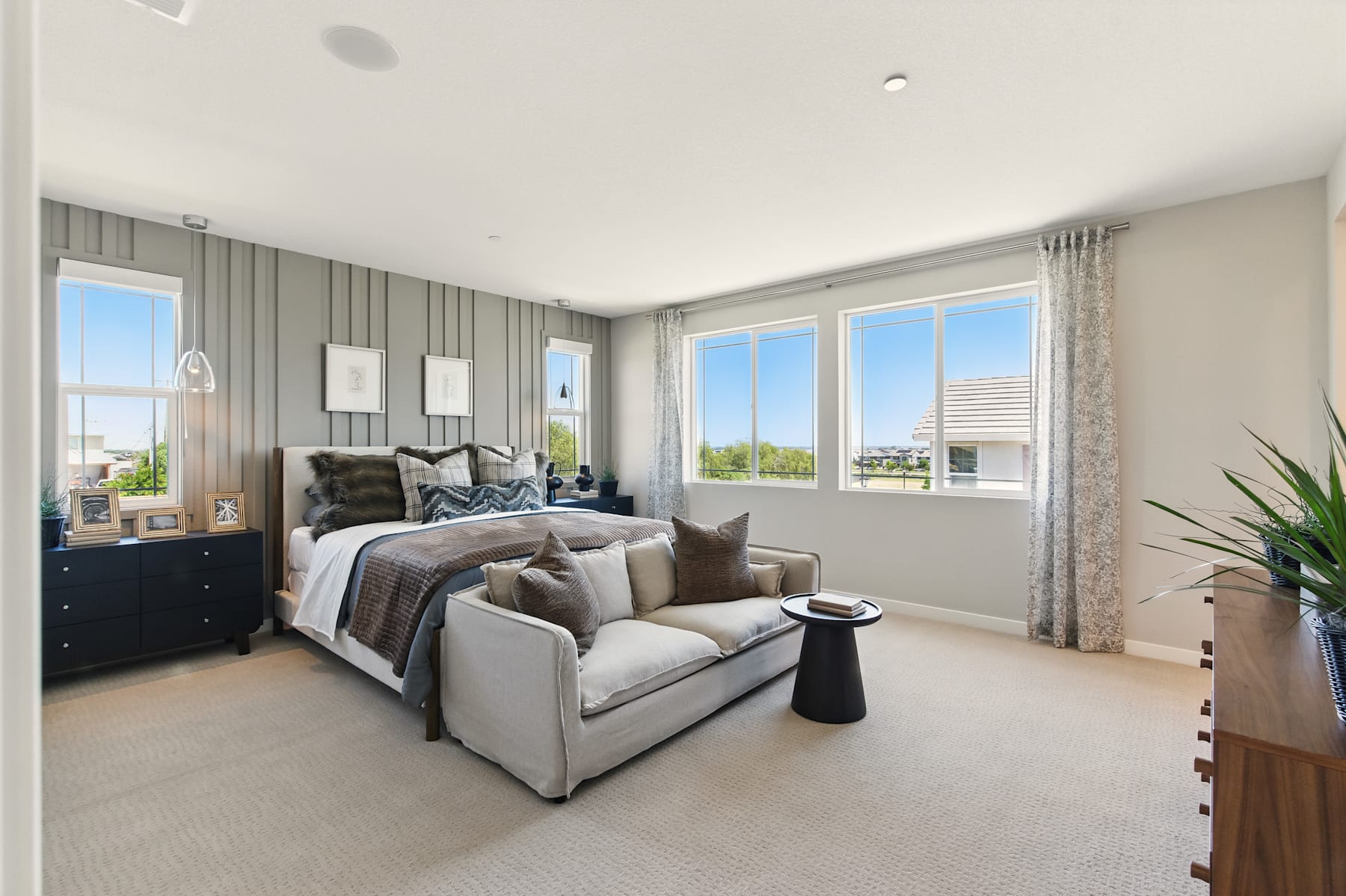 A cozy and modern bedroom with a large window overlooking a scenic outdoor view, featuring a plush gray sofa and a well-decorated bed with patterned bedding.