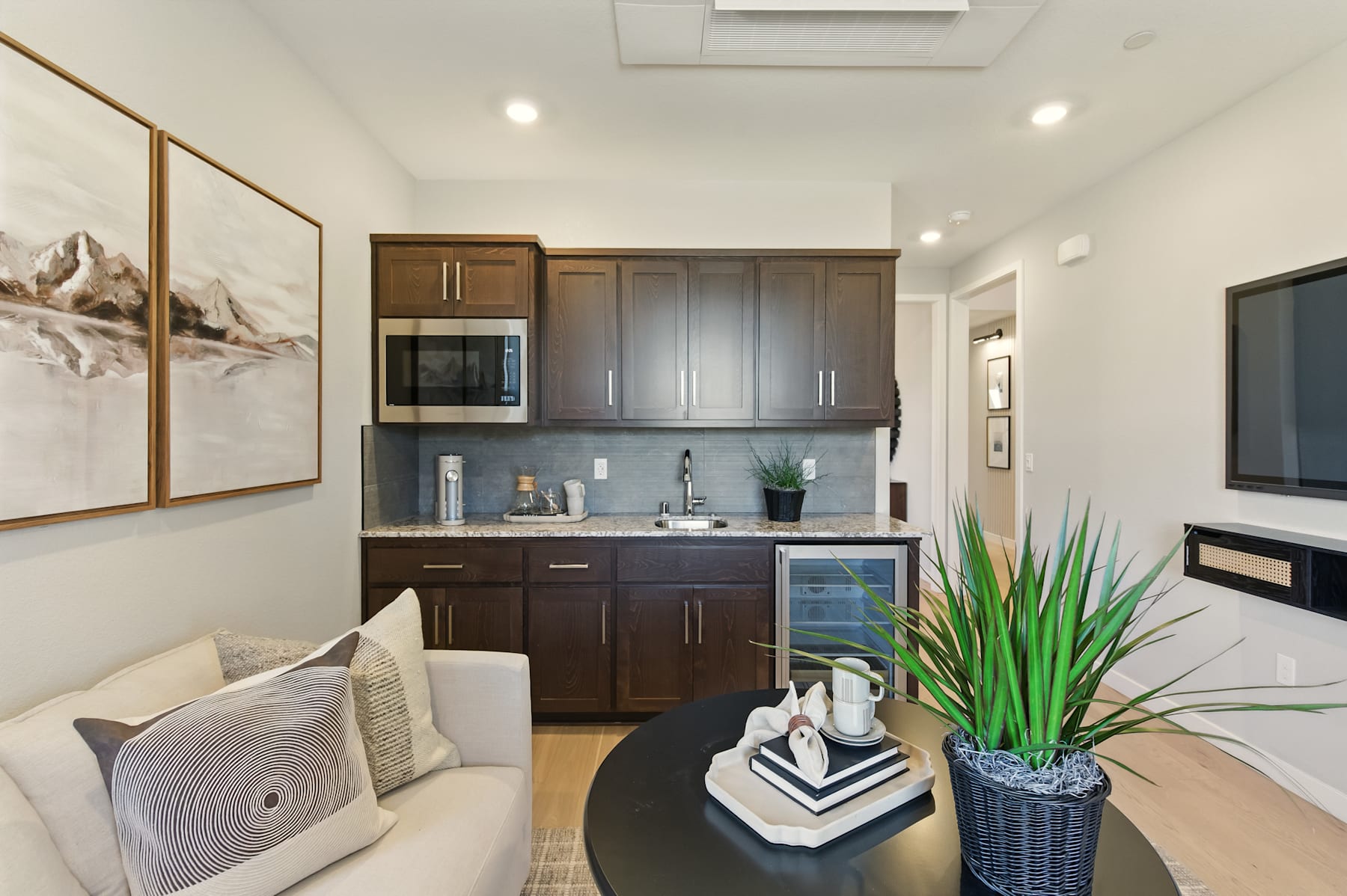 A modern and well-designed kitchen area with dark wood cabinets, a white backsplash, and a large framed artwork on the wall, complemented by a cozy living space with a white sofa and a potted plant.