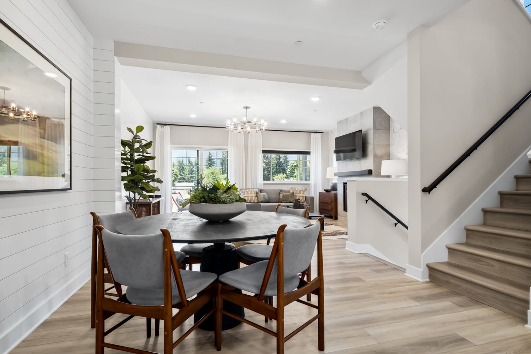 A modern and bright dining area with a round wooden table and chairs, surrounded by a spacious living room with large windows and a staircase leading to the upper level.