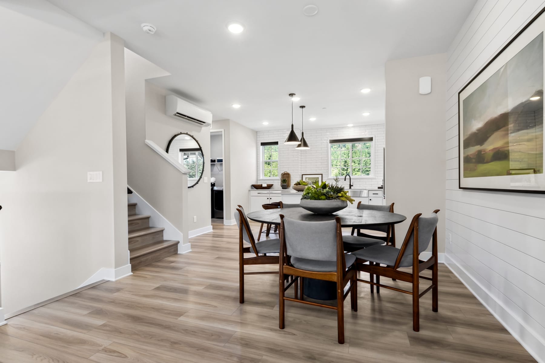 A modern and minimalist dining area with a round wooden table surrounded by chairs, set against a bright and airy backdrop with white walls, hardwood floors, and a large window overlooking greenery.