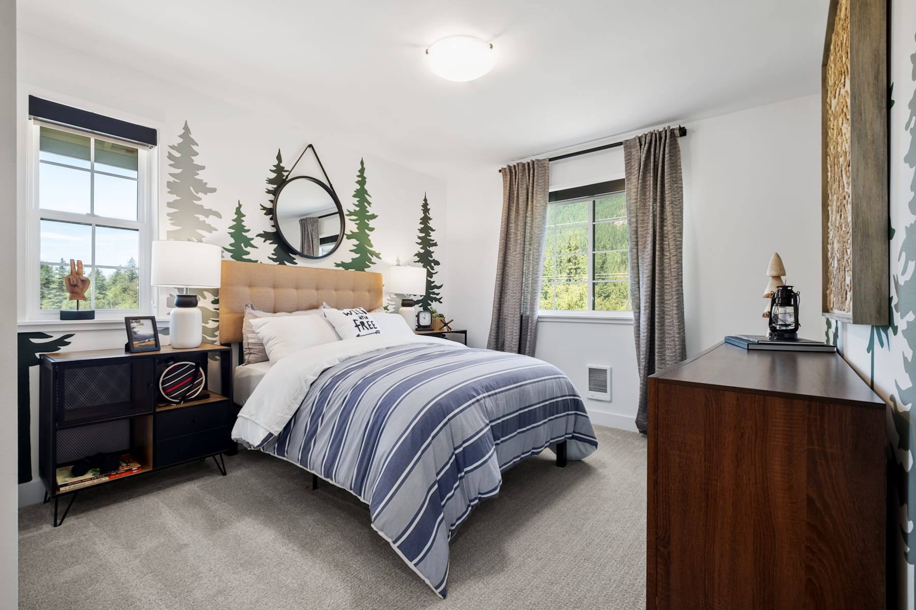 A cozy and rustic bedroom with a large window overlooking a forested landscape, featuring a plush bed with a striped comforter, a wooden nightstand, and decorative elements like a round mirror and wall art depicting pine trees.