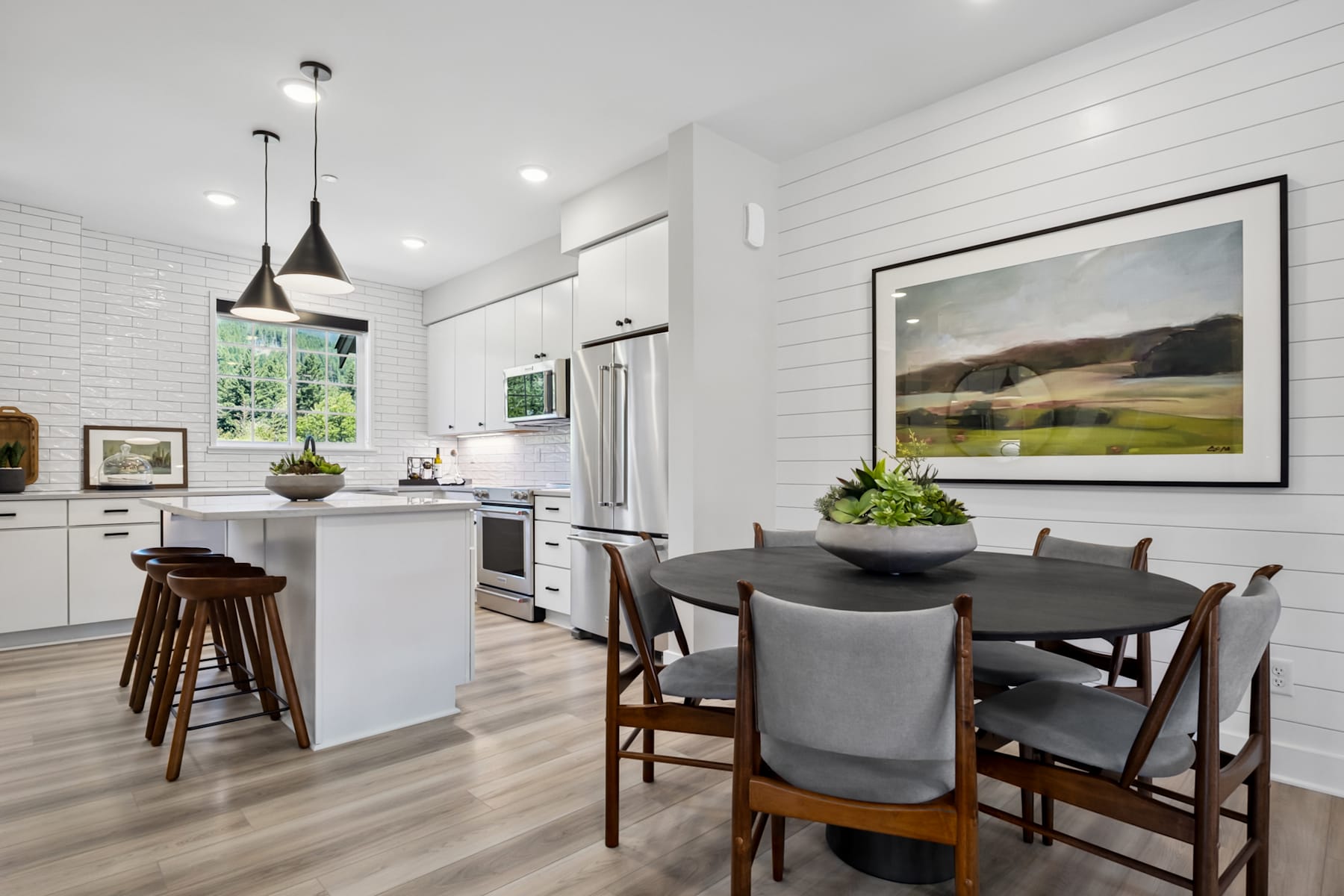 A modern, open-concept kitchen and dining area with white walls, wooden floors, and a large framed landscape painting on the wall.