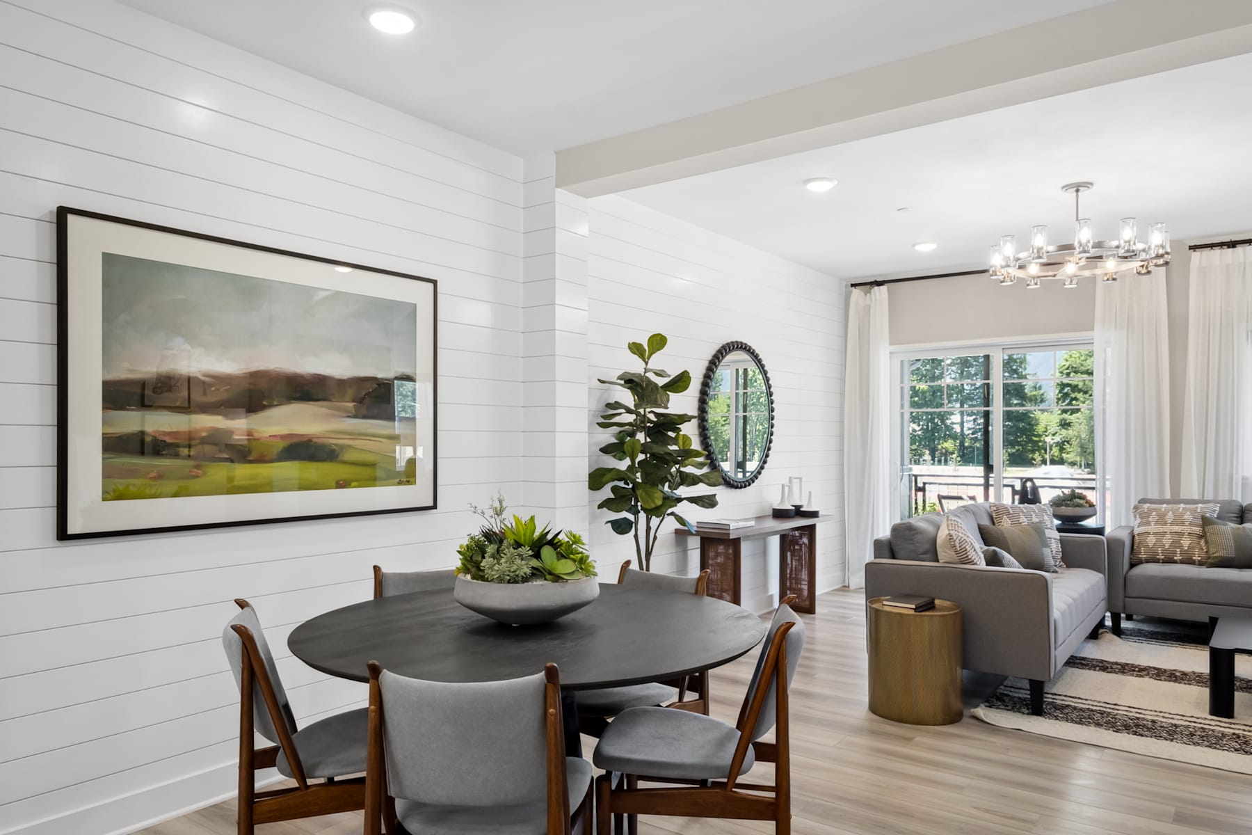 A cozy and inviting dining area with a round wooden table and gray chairs, surrounded by a bright and airy living room with a large landscape painting, a mirror, and various decorative elements.