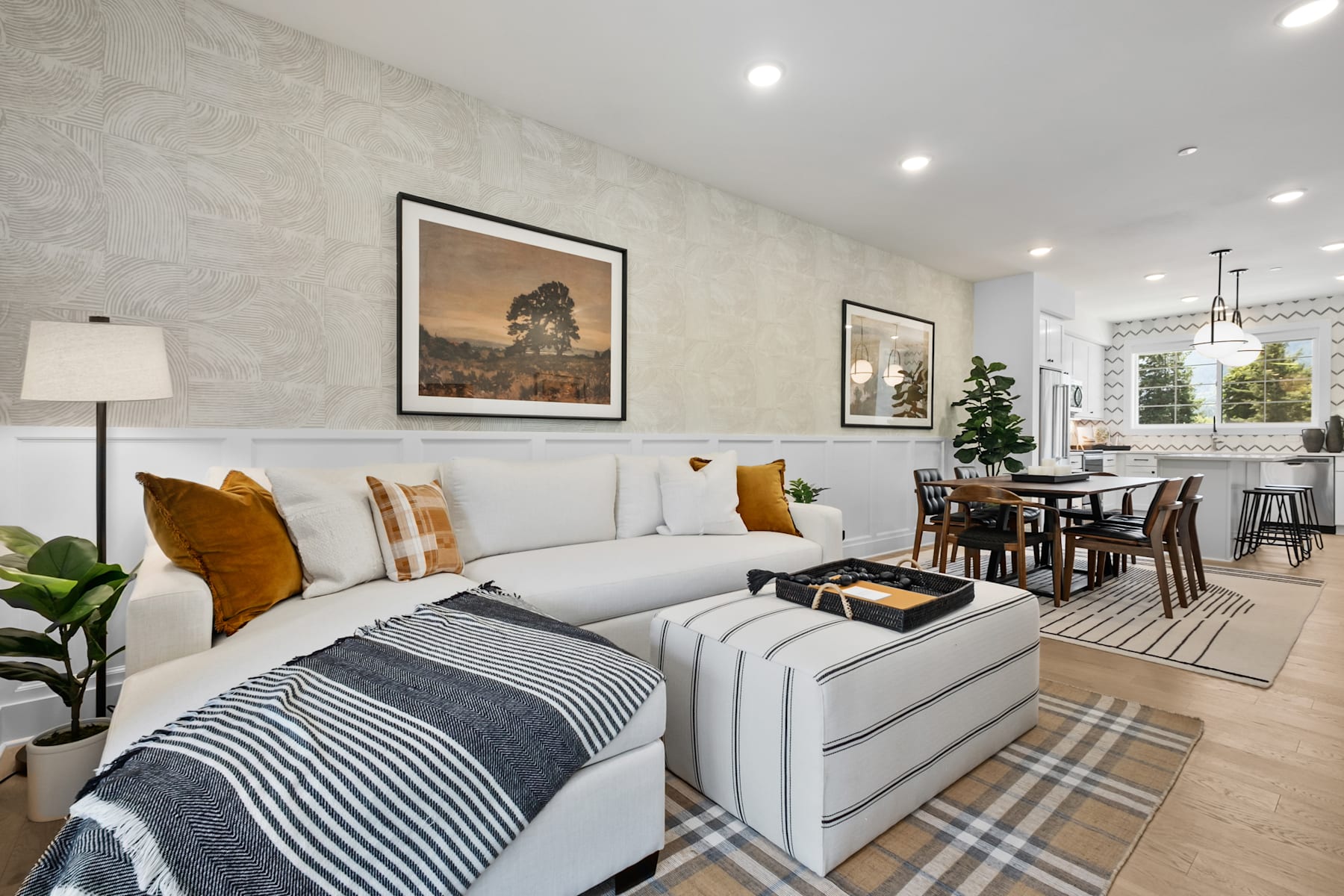 A cozy and inviting living room with a white sofa, patterned throw pillows, and framed artwork on the walls, leading into a dining area with a wooden table and chairs.