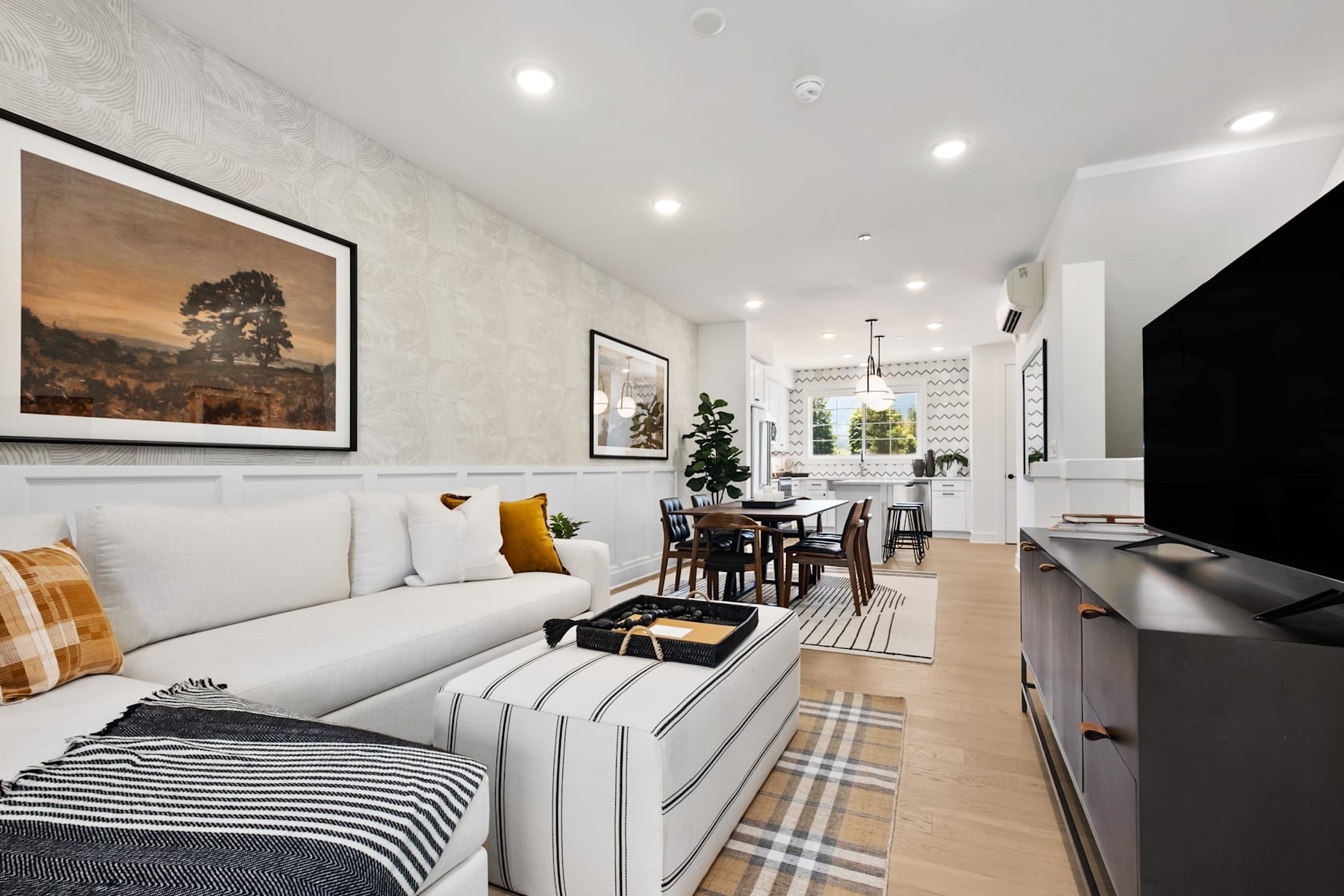 A cozy and modern living room with a white sofa, a striped rug, and framed artwork on the walls, leading into a dining area with a wooden table and chairs.