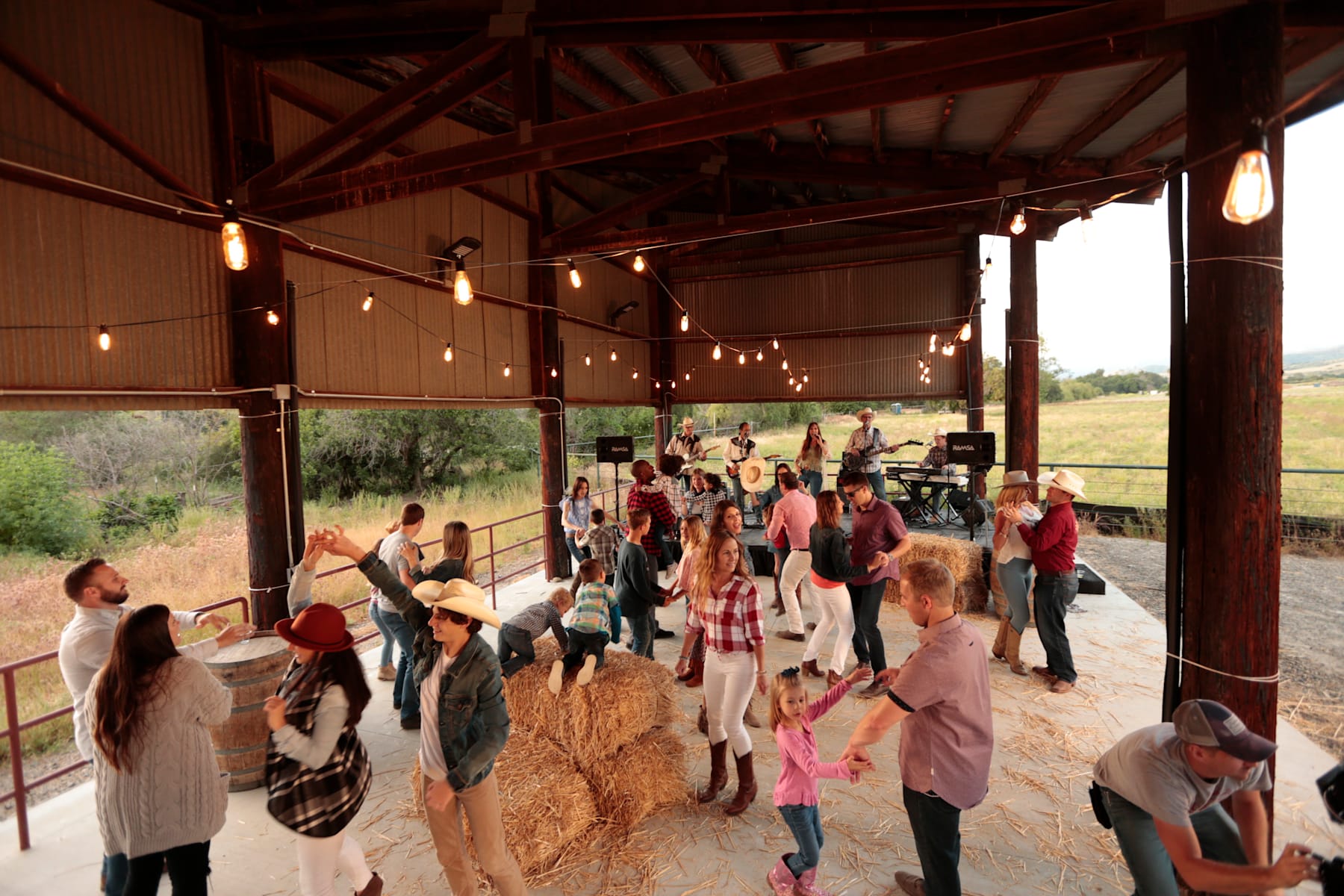 A crowded outdoor gathering with people mingling under a covered porch, surrounded by a rural landscape.