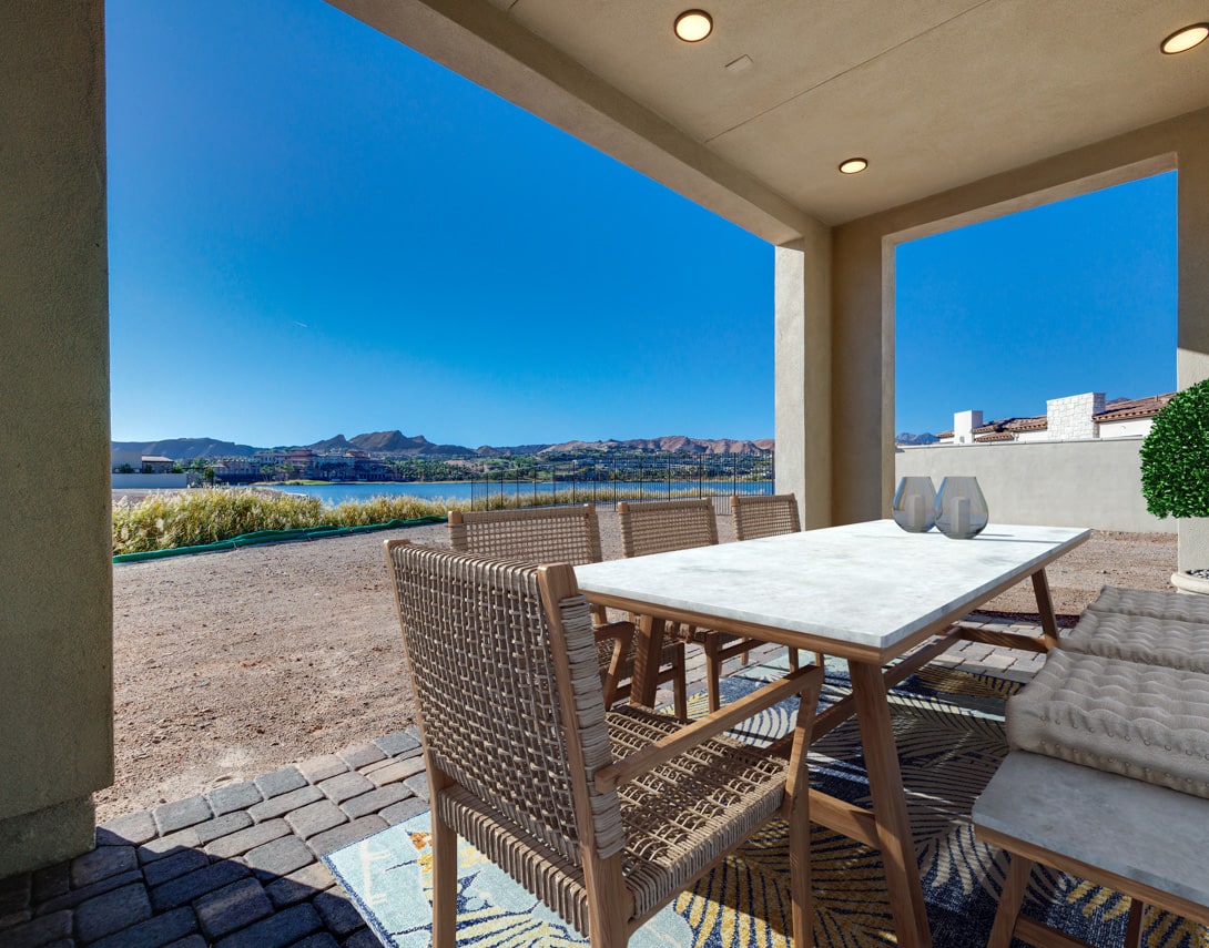 A modern outdoor dining area with a wooden table and wicker chairs, overlooking a scenic waterfront landscape with mountains in the background.