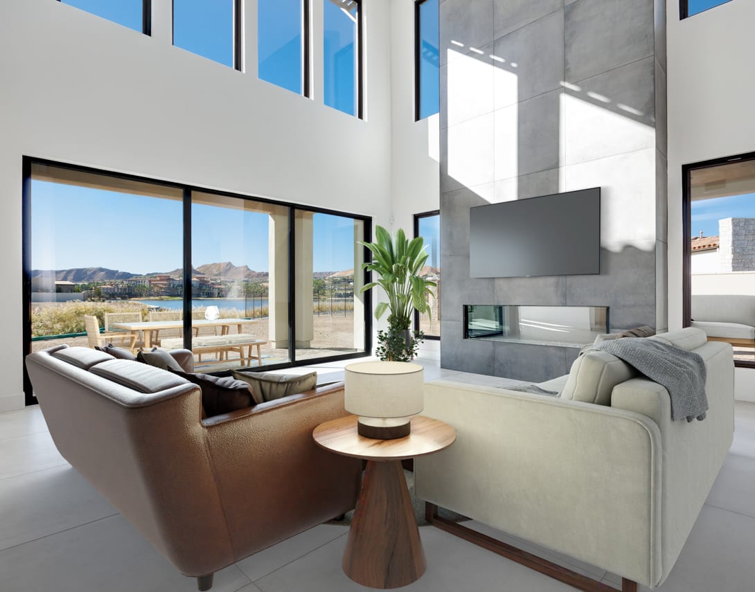A modern and spacious living room with large windows overlooking a scenic outdoor landscape, featuring comfortable sofas, a coffee table, and a potted plant.