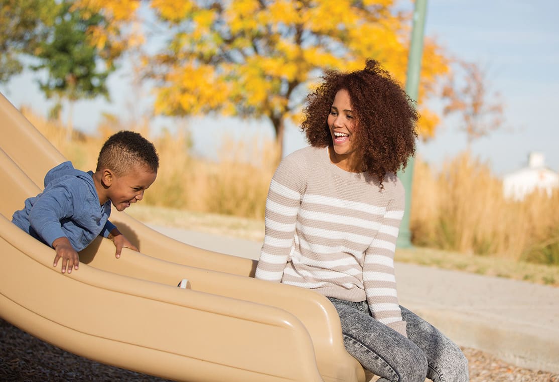 A young woman with curly hair is smiling and interacting with a young boy on a playground slide in an autumn setting with yellow trees in the background.