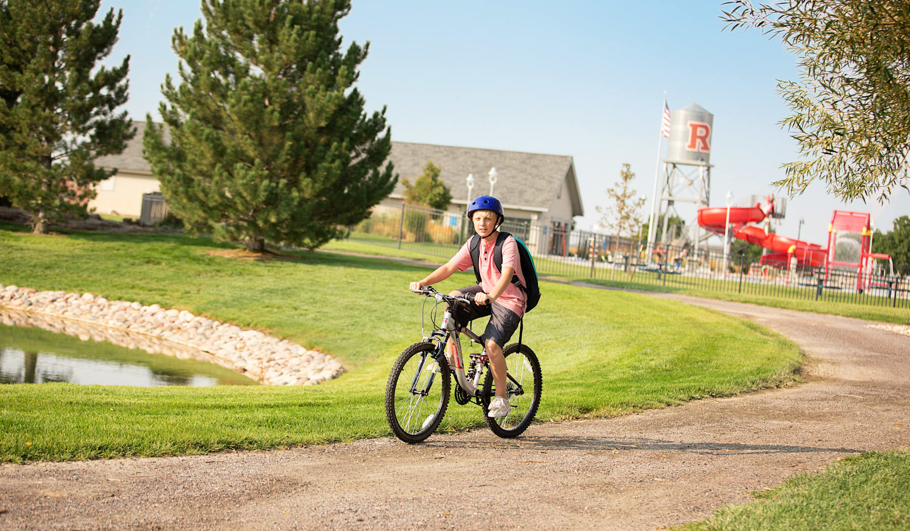 A person wearing a blue hat and a pink jacket is riding a bicycle on a dirt path surrounded by lush greenery, with buildings and signage visible in the background.