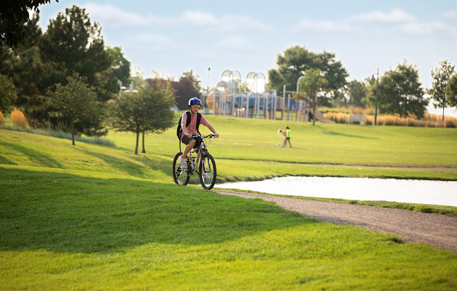 A person on a bicycle rides along a paved path through a lush, green park with trees and playground equipment visible in the background.