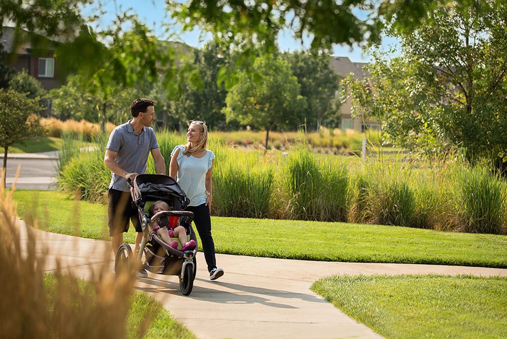 A young couple strolling along a paved path in a lush, green park, with a stroller in tow, surrounded by trees and a well-manicured lawn.