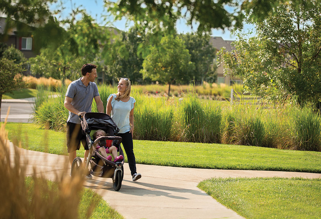 A couple walks together on a paved path, pushing a stroller, surrounded by lush greenery and a well-manicured lawn in the background.
