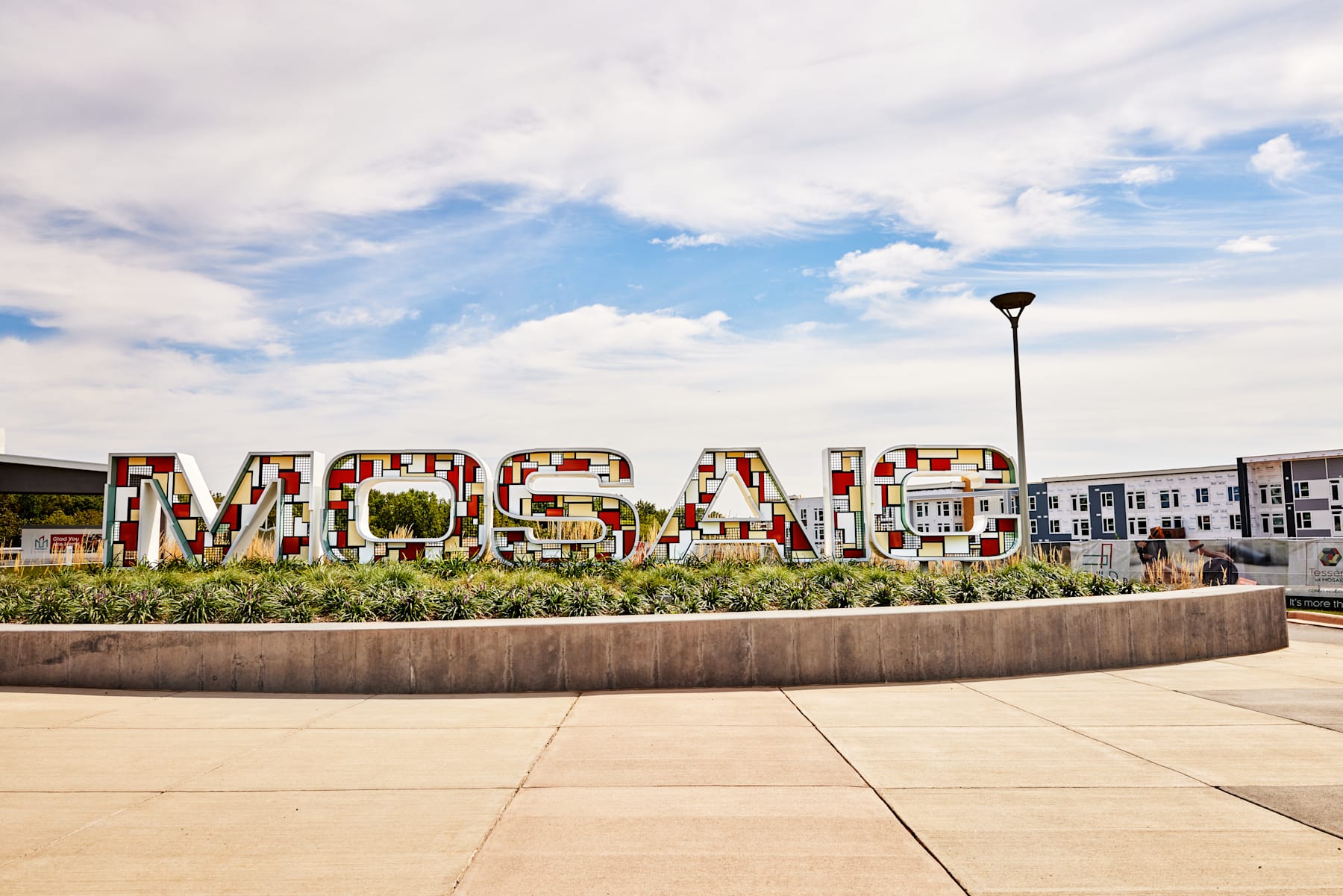 A large, colorful "Mosaic" sign stands in the foreground, surrounded by a paved plaza and landscaping, with a cloudy blue sky and buildings visible in the background.