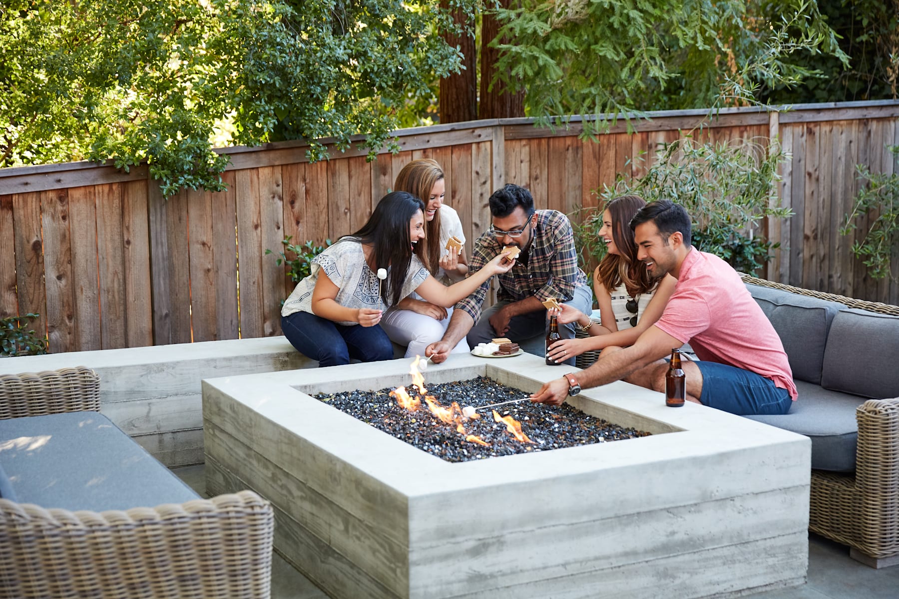 A group of friends gathered around a concrete fire pit, enjoying each other's company in a cozy outdoor setting surrounded by lush greenery and a wooden fence.