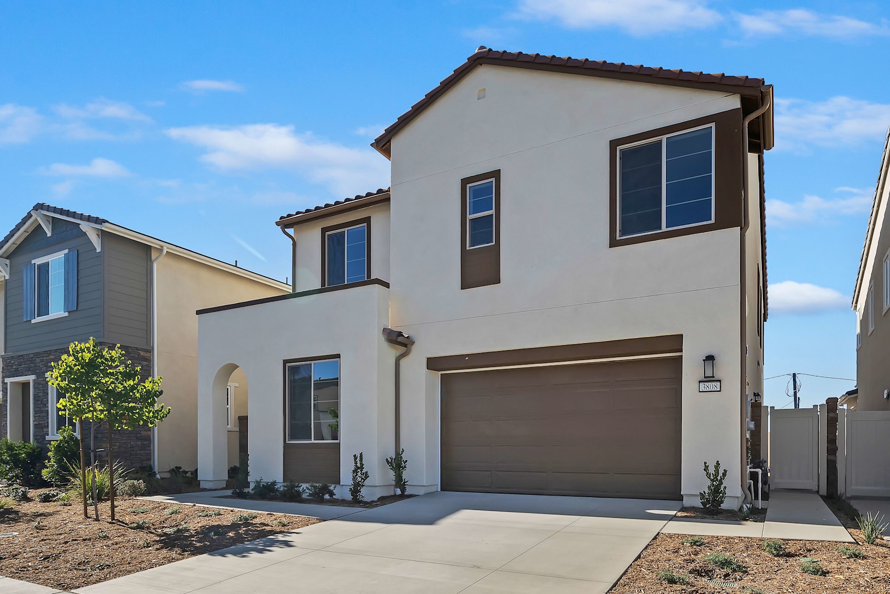 A residential neighborhood with modern, two-story houses, a paved driveway, and landscaped yards in the foreground, set against a clear blue sky in the background.