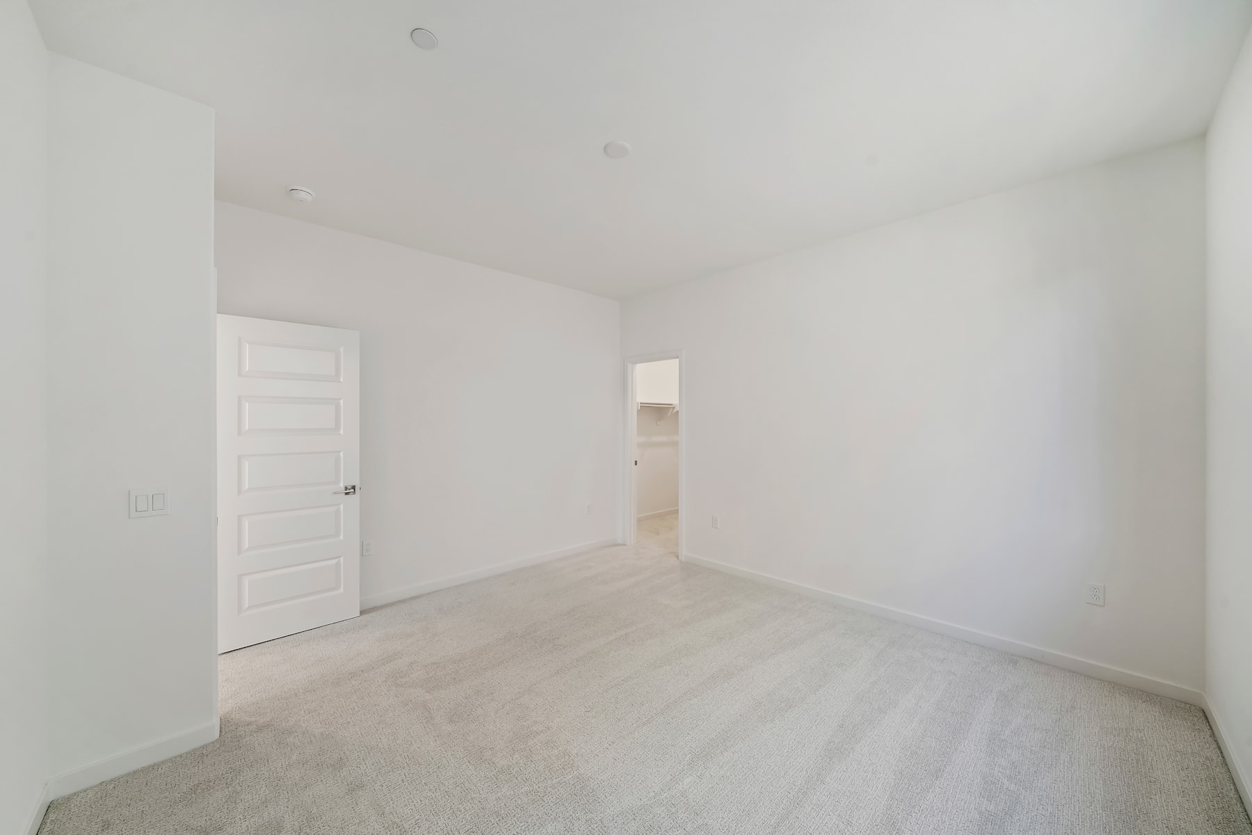 An empty, white-walled room with a hardwood floor and a closed door visible in the background.