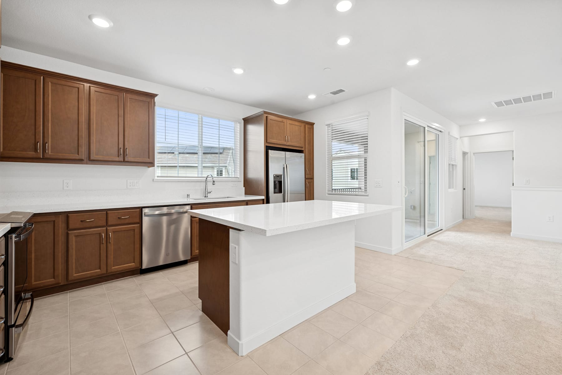 A modern, open-concept kitchen with wooden cabinets, white countertops, and a central island, set against a bright and airy background with large windows.