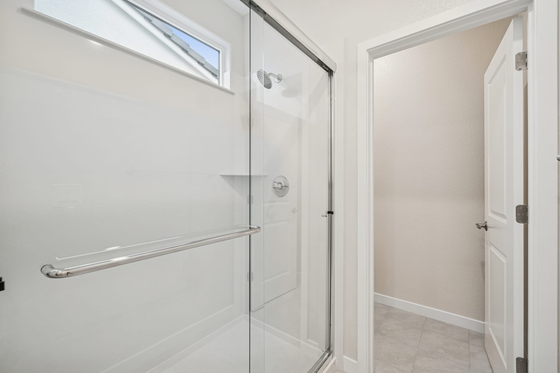 A modern, minimalist bathroom with a glass shower enclosure, a white door, and a hallway visible in the background.