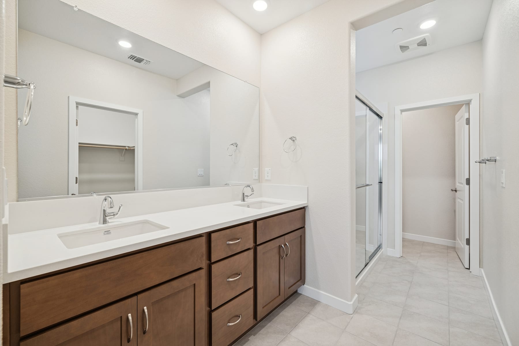 A modern and spacious bathroom with a large vanity, double sinks, and a tiled floor. The room is well-lit and features a hallway leading to other areas of the home.