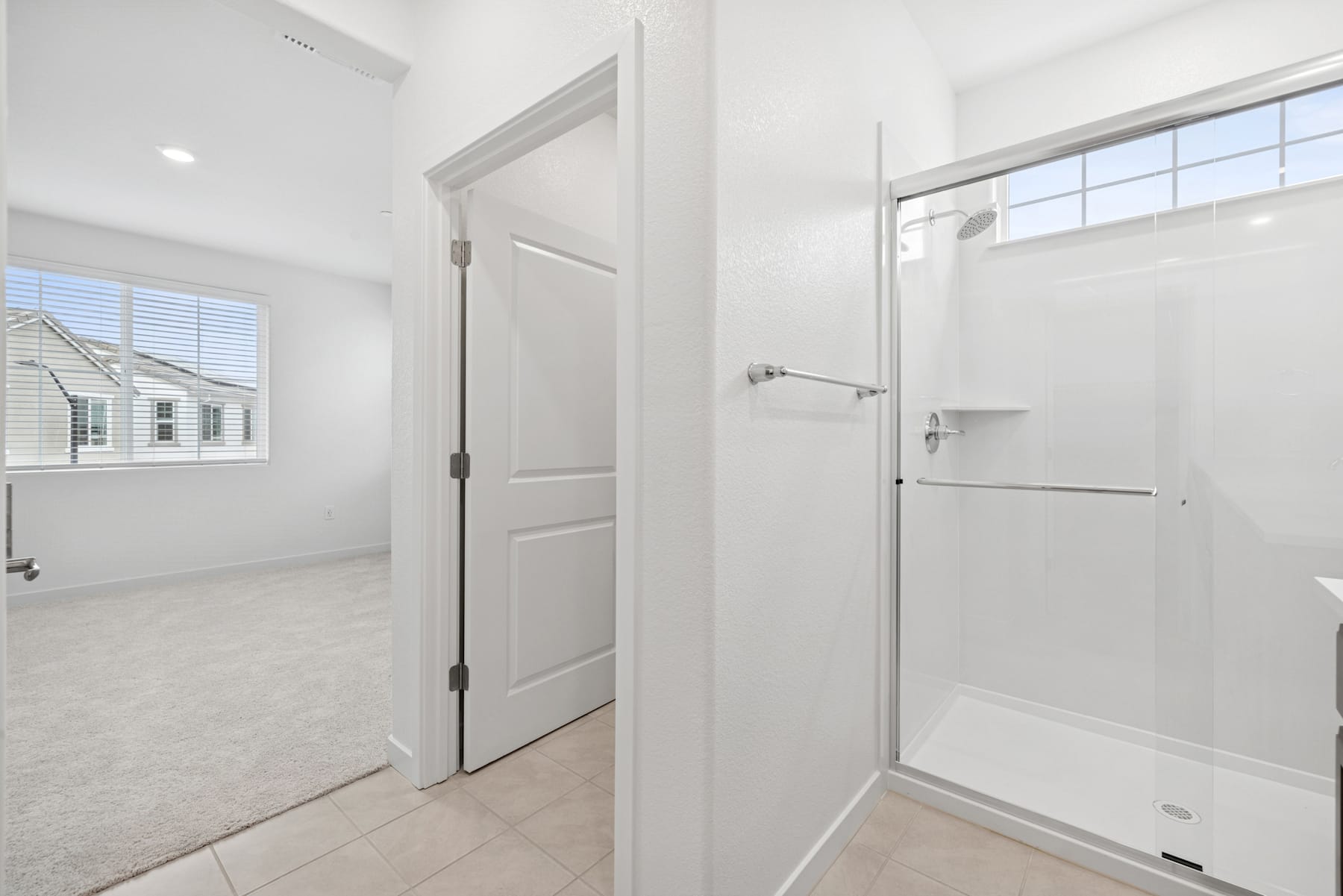 A bright, minimalist bathroom with a white door, shower, and window overlooking a residential neighborhood.
