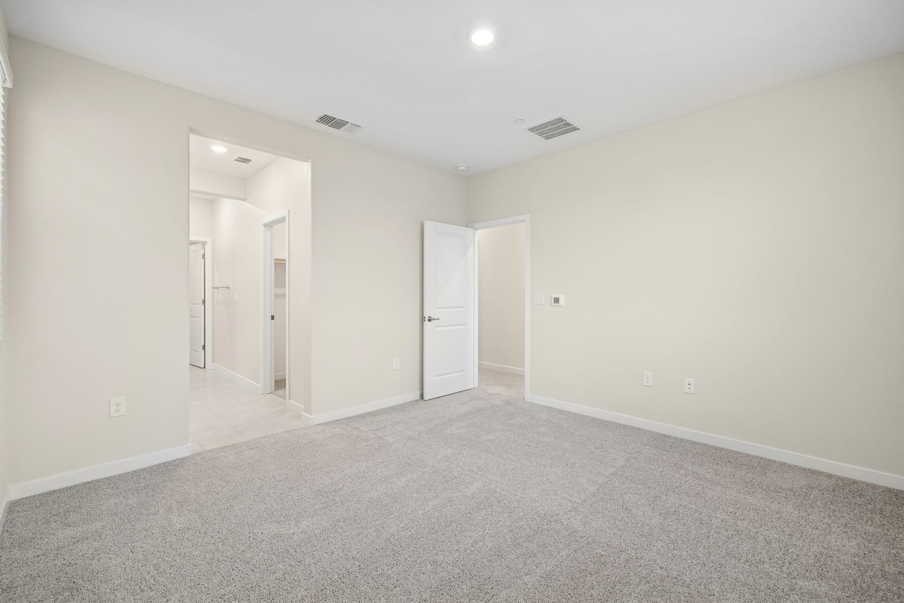 An empty, well-lit room with white walls, a gray carpeted floor, and two closed doors leading to other areas.