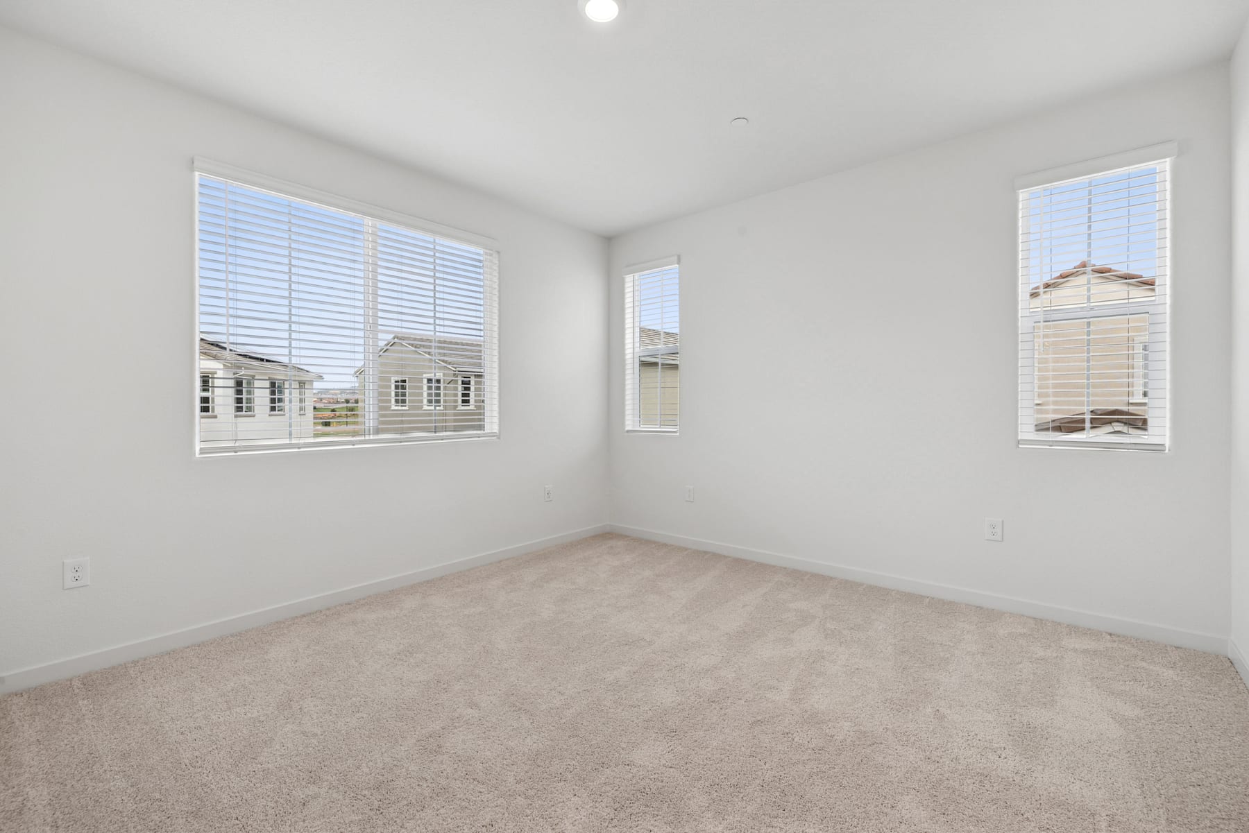 A spacious, well-lit room with white walls, carpeted floor, and large windows providing a view of the exterior buildings.