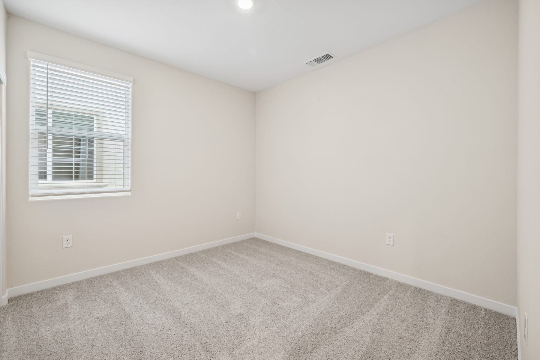 A simple, minimalist bedroom with a window, white walls, and gray carpeted flooring.