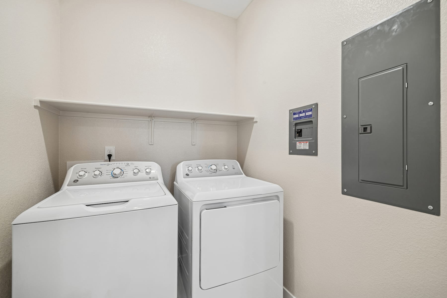 A small laundry room with a washing machine and dryer, along with a control panel and a door in the background.