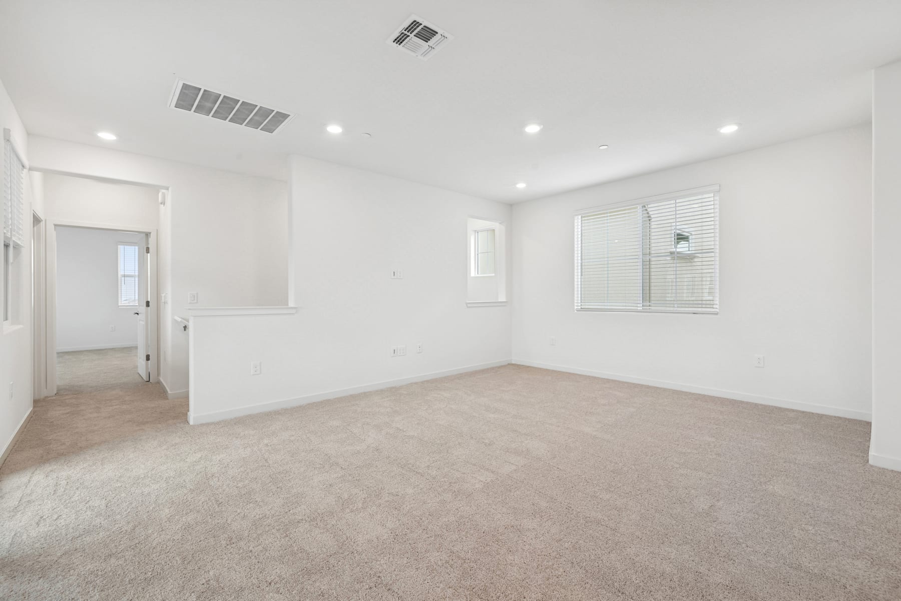 A spacious, well-lit room with white walls, a carpeted floor, and several recessed ceiling lights. The room appears to be a bedroom or living space, with a window visible in the background.