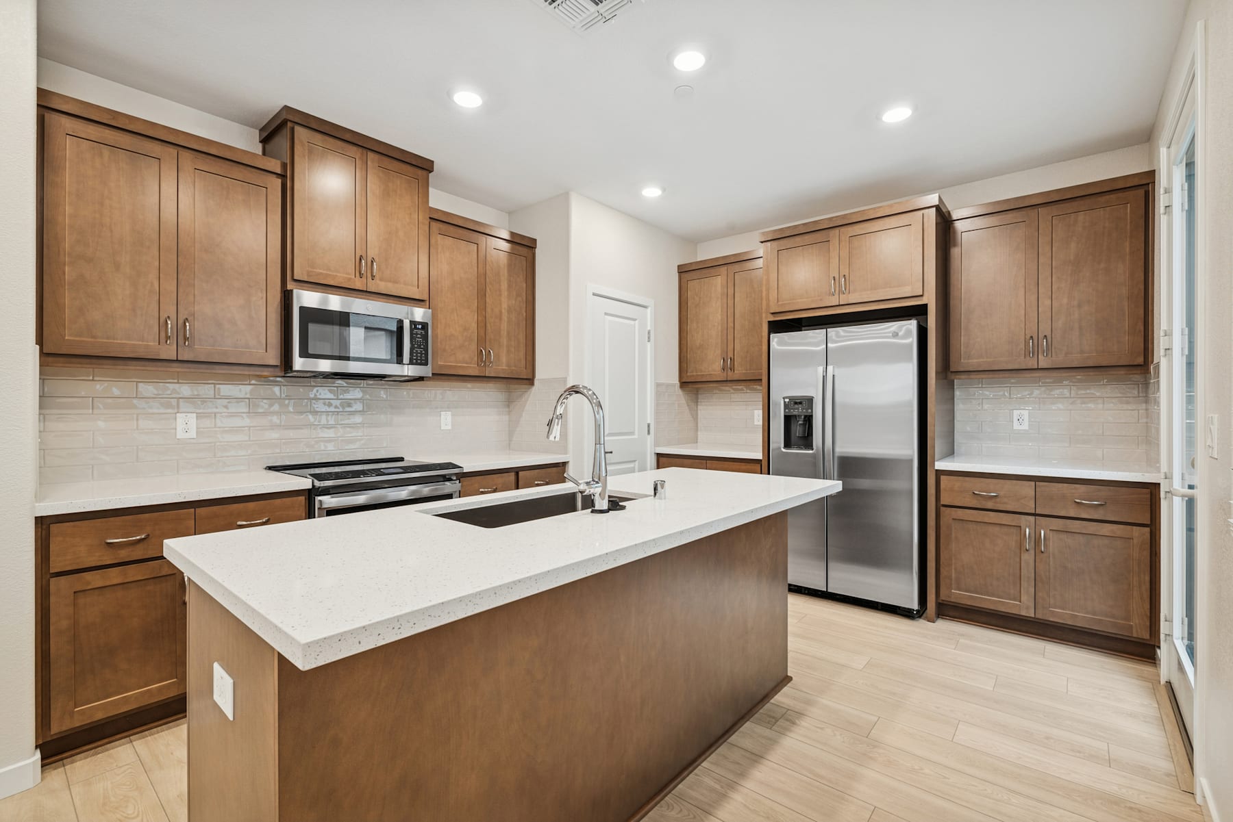 A modern kitchen with wooden cabinets, a white countertop, and stainless steel appliances, set against a light-colored hardwood floor.